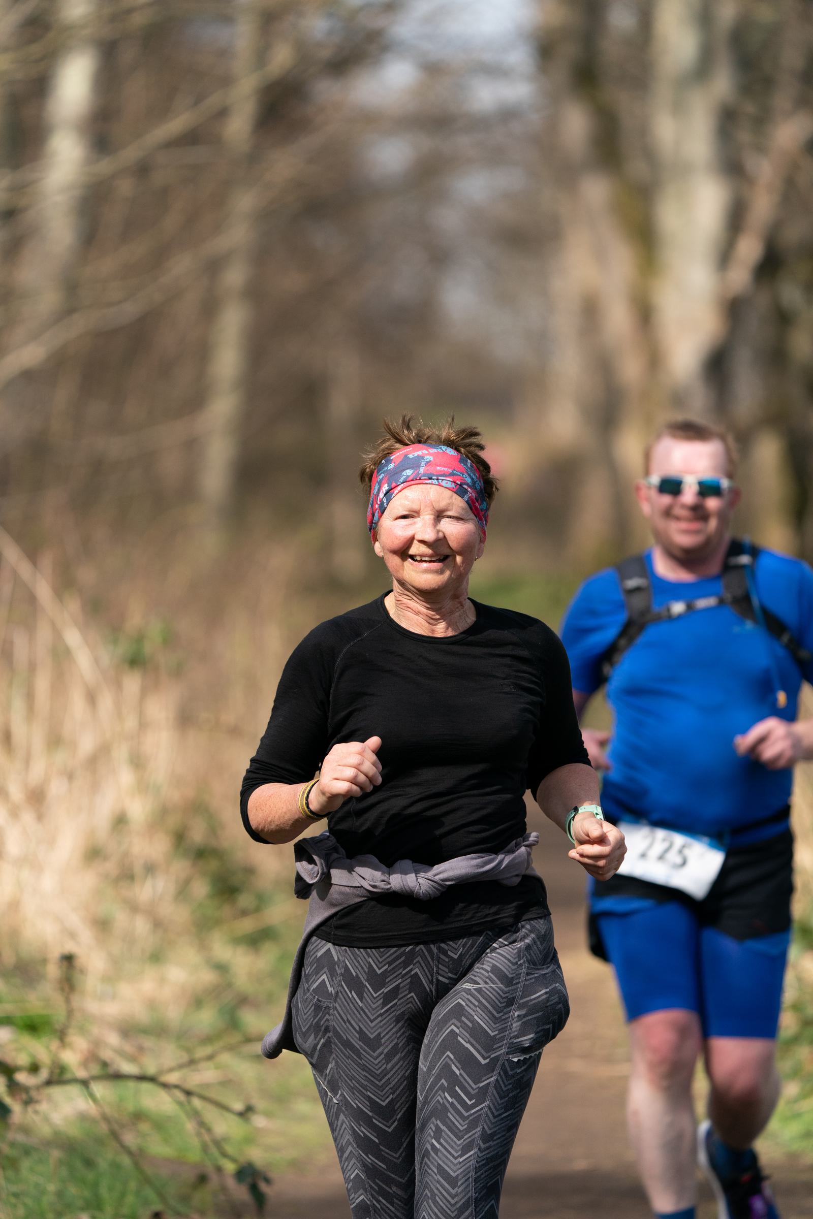 A woman runs on a wooded trail wearing a black top and patterned grey pants, smiling and wearing a colorful headband. Behind her, a man in blue athletic gear with a race bib also runs. The weather appears mild and the trees are bare.