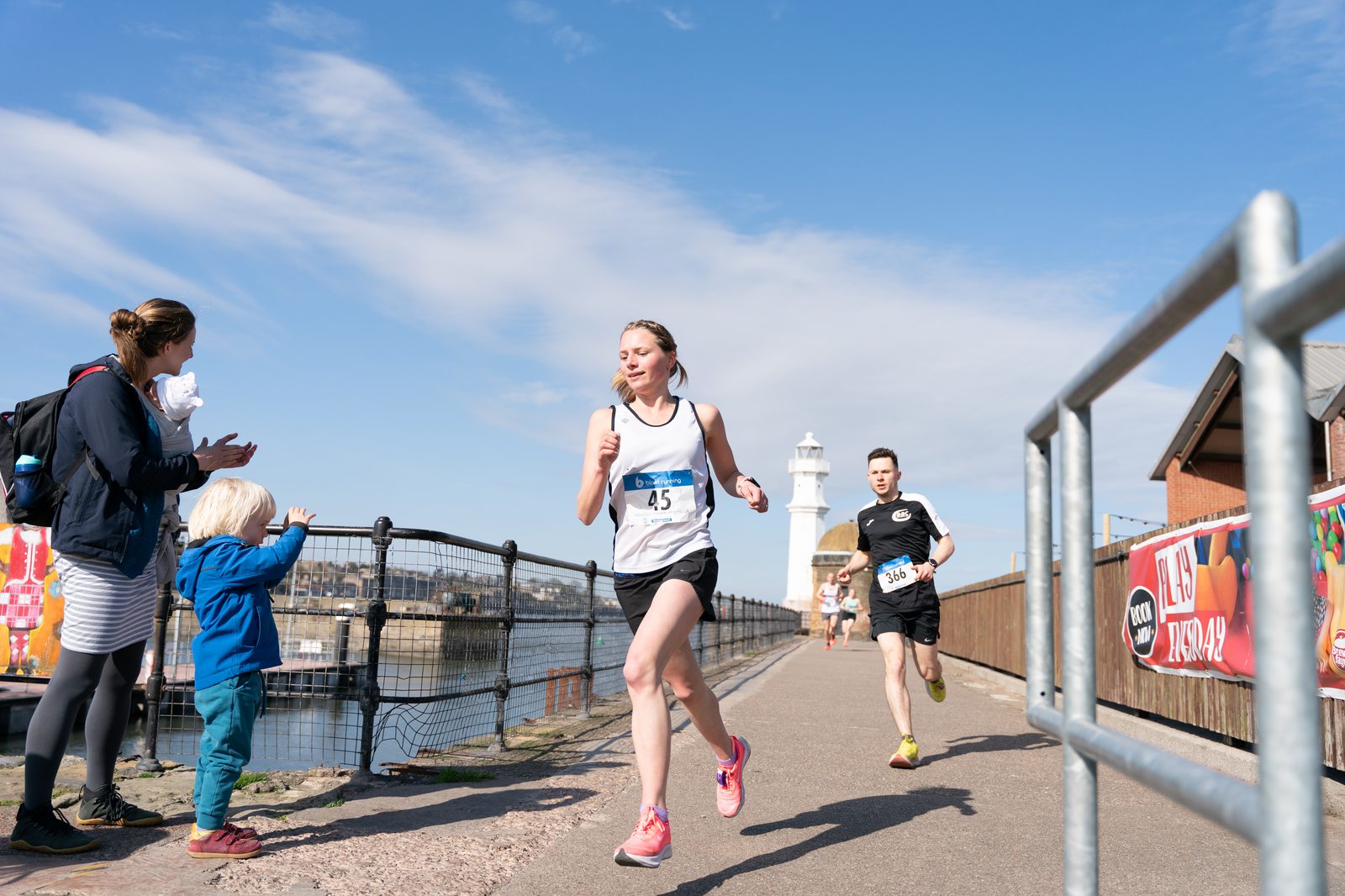 A woman and a man are running a race on a scenic coastal pathway with a lighthouse in the background. A woman and a child stand by the sideline, clapping and cheering. The sky is clear and blue, and a metal railing lines the path.