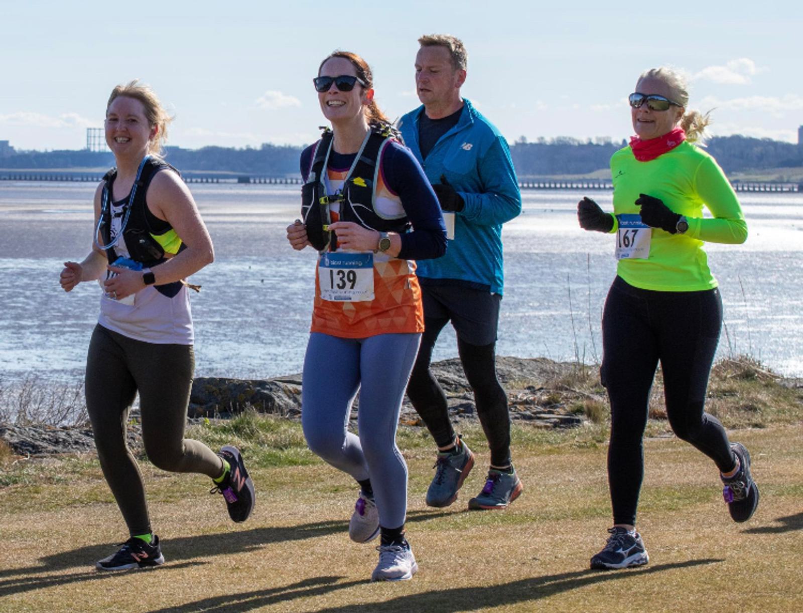 Four people are jogging along a waterfront on a sunny day. They are dressed in athletic gear, wearing numbers, and look happy. In the background, there's a body of water and a distant shoreline with trees and buildings.