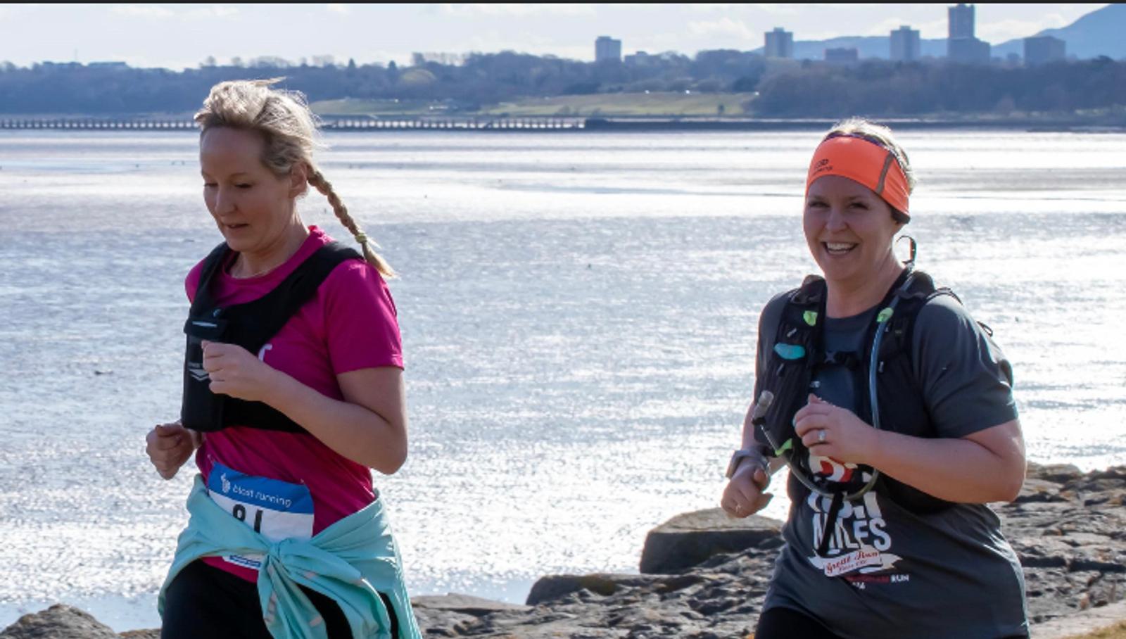 Two women are running on a rocky coastline path. The woman on the left, wearing a pink shirt, focuses ahead, while the woman on the right, wearing an orange headband and gray shirt, smiles. The background shows a body of water and distant buildings.