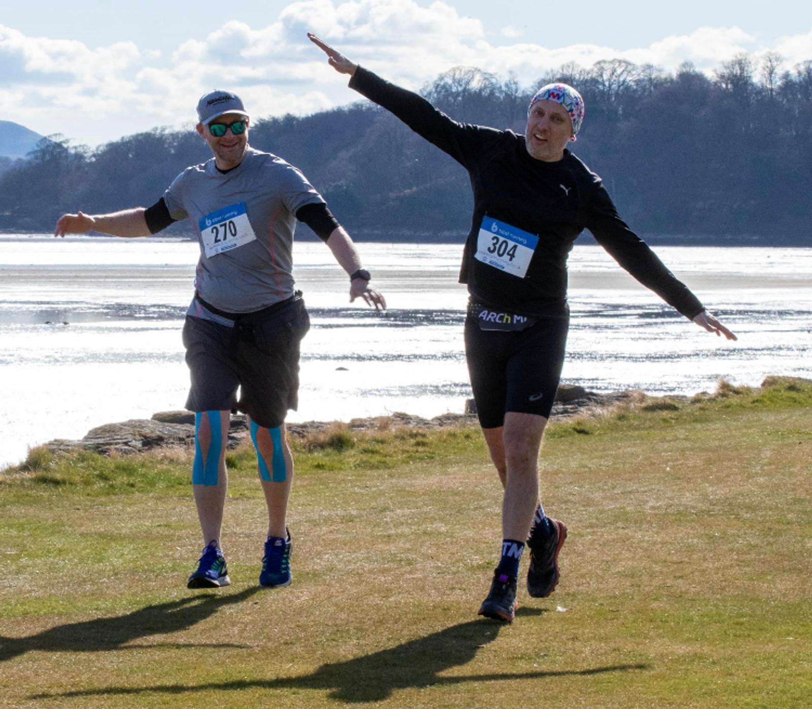 Two smiling runners are enjoying a sunny run on a grassy path alongside a water body. One runner (number 270) has blue knee tape and blue shoes, while the other (number 304) wears a colorful bandana and extends his arms playfully. The background is scenic with trees and hills.