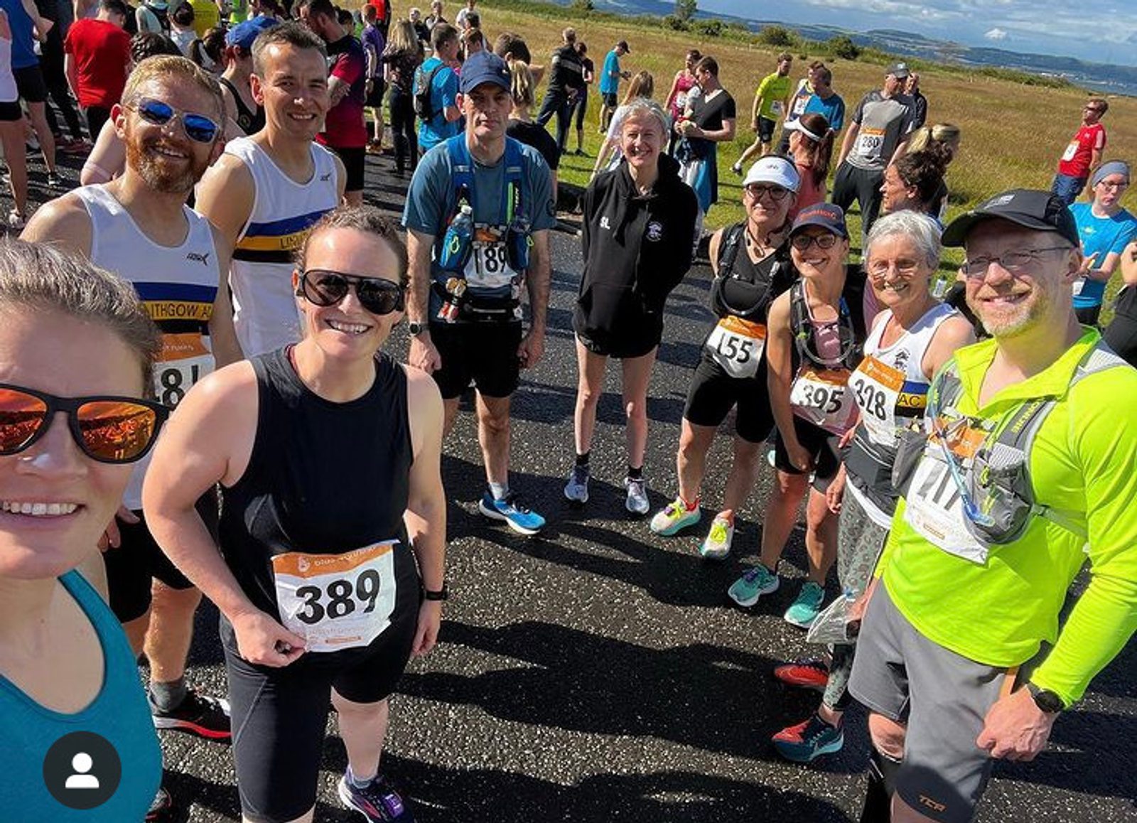 A group of runners in race attire stands together on a road for a photo, each wearing a numbered bib. They smile and pose for the camera with a scenic landscape of fields and a distant horizon behind them. The weather is sunny and clear.