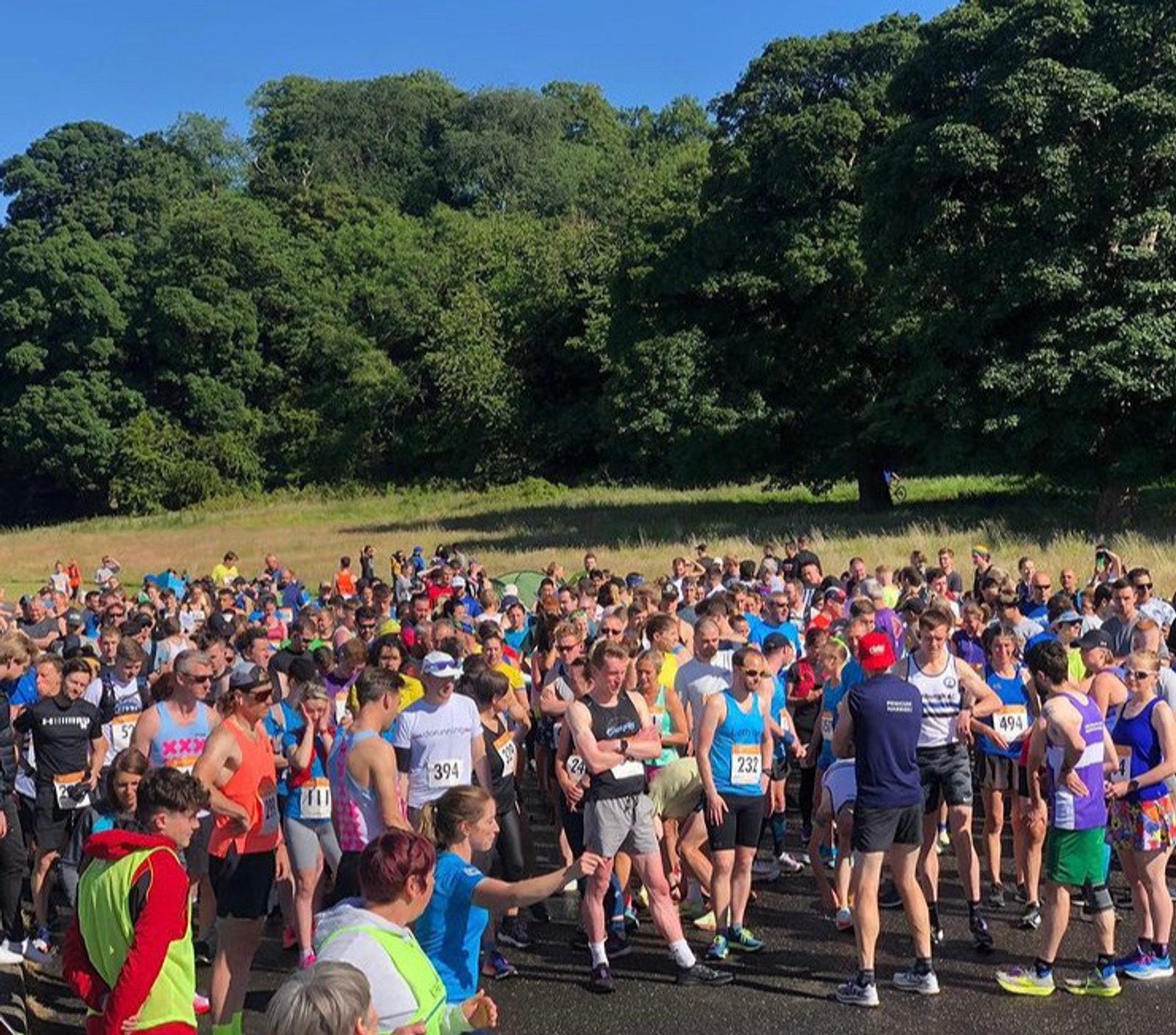 A large group of runners gather at the starting line of an outdoor race. Many are dressed in brightly colored athletic gear. The background features lush green trees and a clear, sunny sky. A few people at the front appear to be giving instructions.