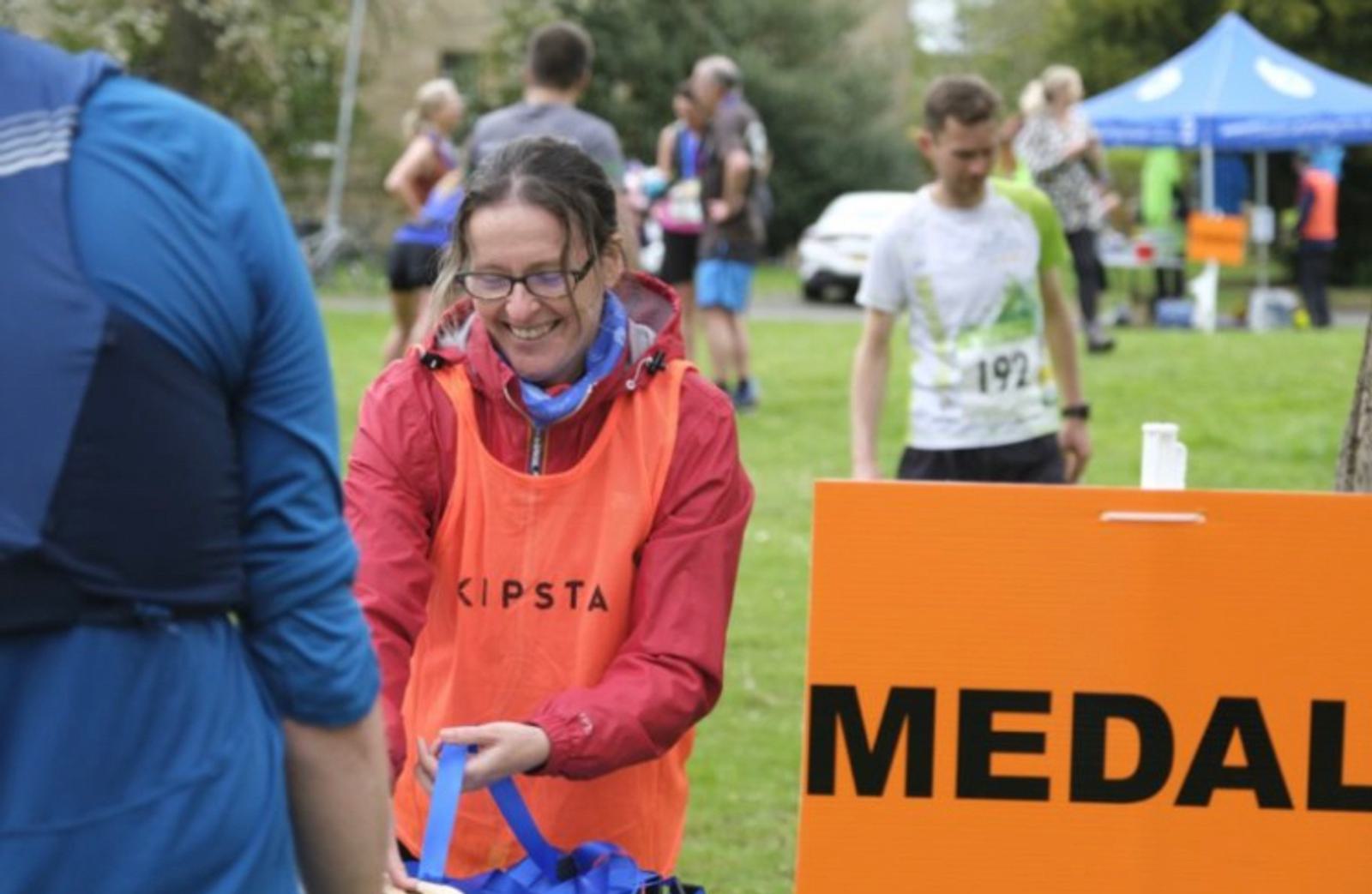 A smiling woman in an orange vest is handing out medals at a race event. Runners in athletic gear are seen in the background. A large orange sign with the word "MEDALS" is visible in the foreground. A blue tent is set up in the background on a grassy area.