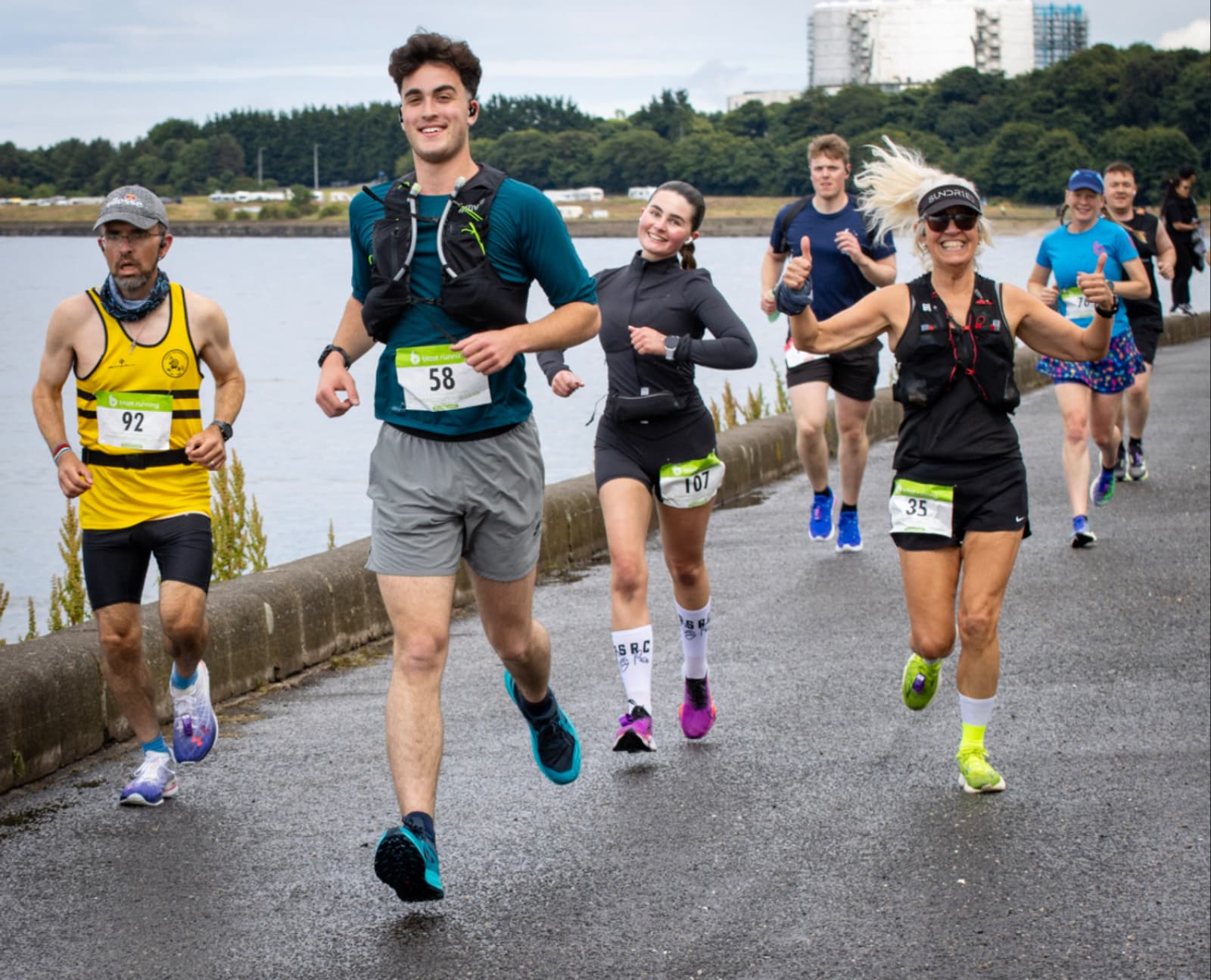 A group of smiling runners, wearing race bibs, jog along a riverside path. Some have their thumbs up and one is mid-jump. The background shows water, greenery, and buildings.