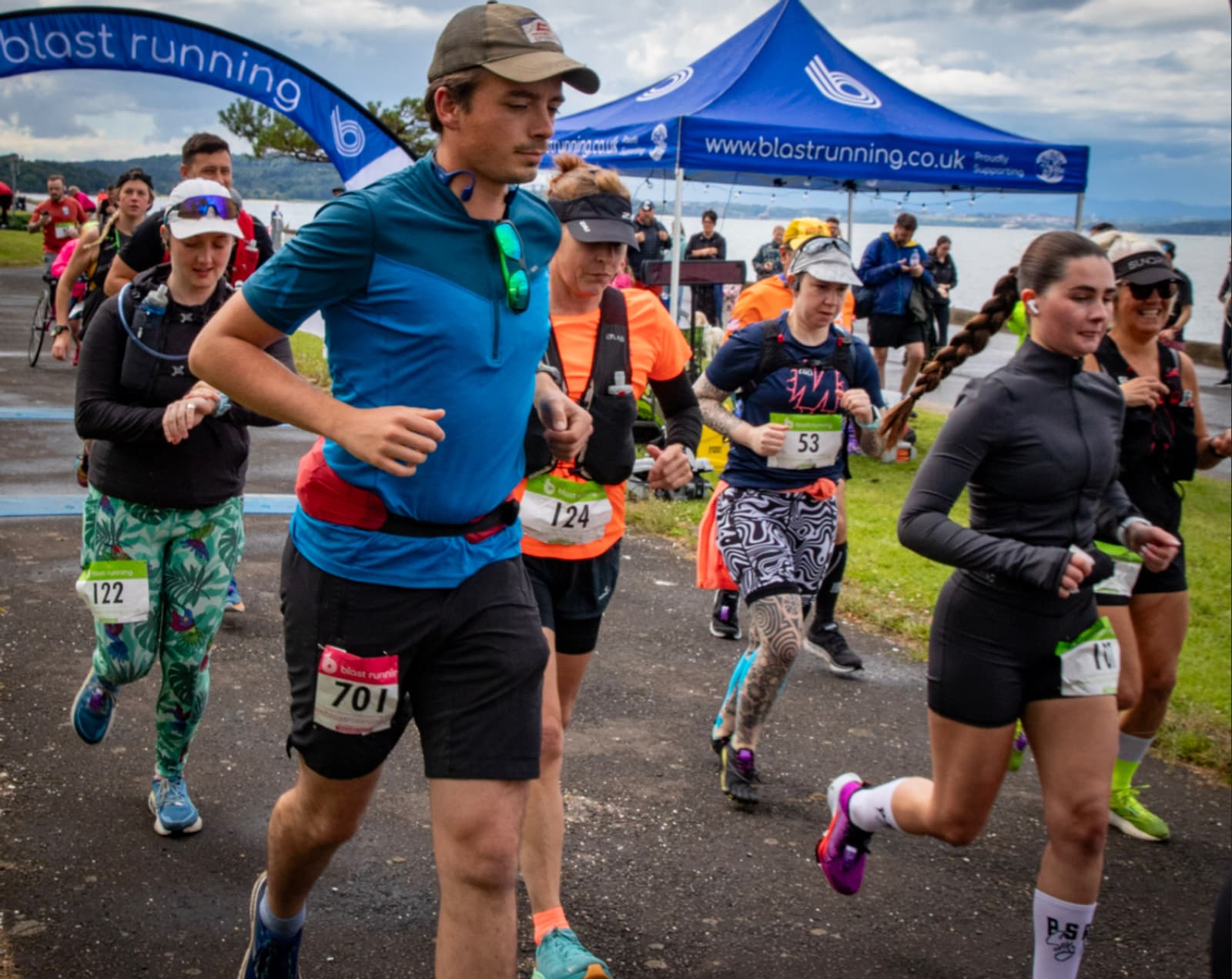 A group of runners wearing race bibs start a race near a blue "blast running" tent by a lakeside on a cloudy day. Some runners wear hats and sunglasses, and trees and water are visible in the background.