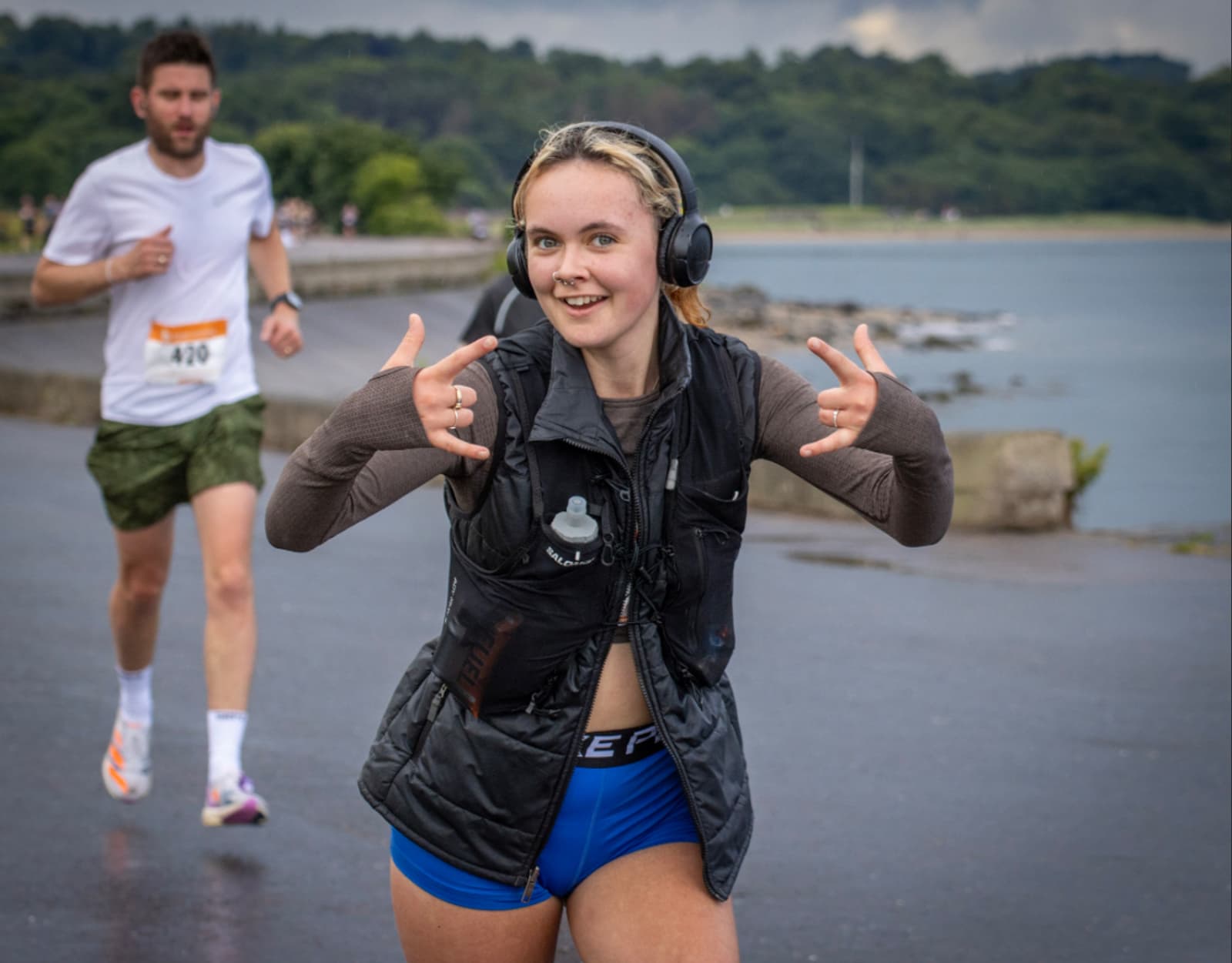 A smiling woman in blue shorts, a vest, and headphones makes hand gestures while running near water, with another runner in the background on a cloudy day.