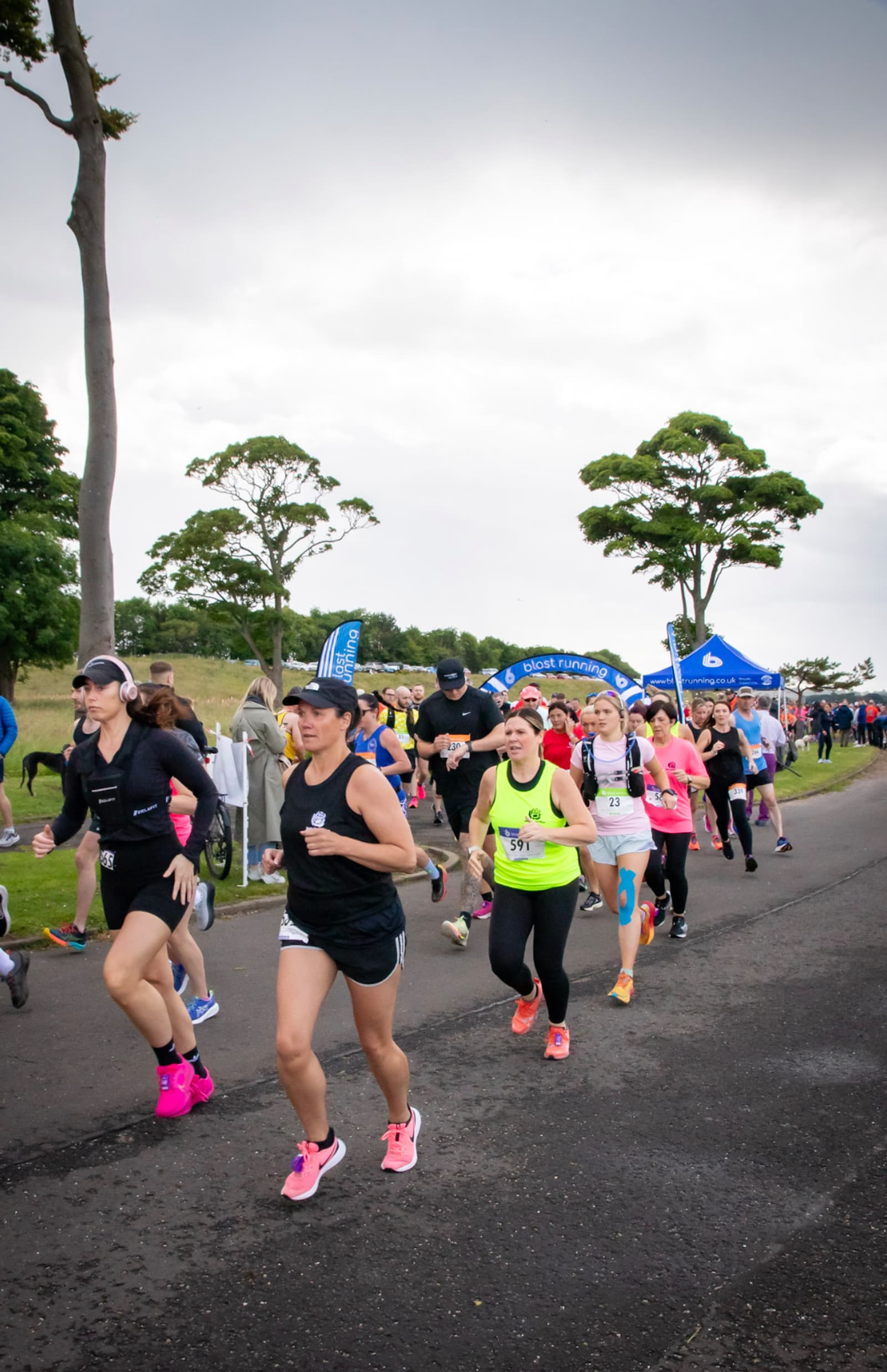 A group of runners in colorful athletic gear participate in a road race on a cloudy day, with trees, banners, and spectators visible in the background.