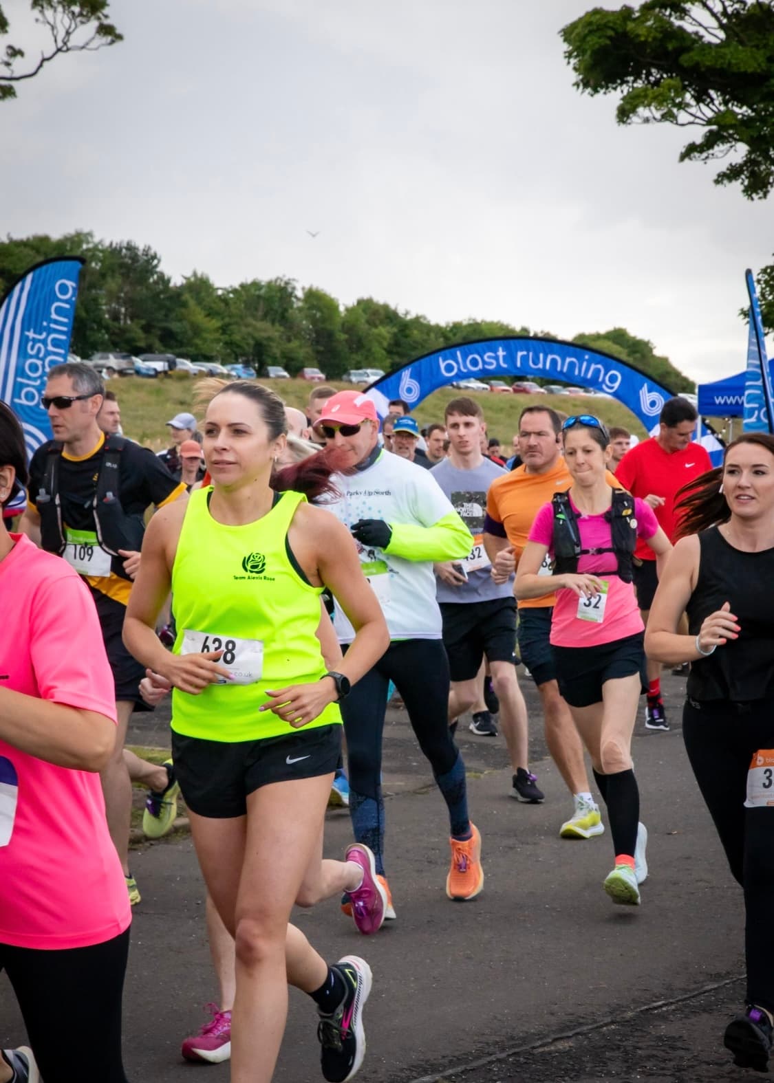 A group of runners with race bibs participate in an outdoor race event, passing under a blue "blast running" banner on a paved road, with trees and parked cars in the background.