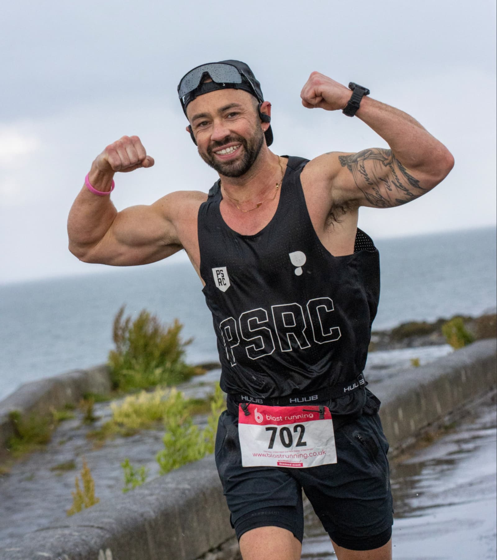 A smiling runner in a black tank top and cap flexes his arms and poses for the camera during a race by the seaside. He wears a race bib numbered 702 and the sea is visible in the background.