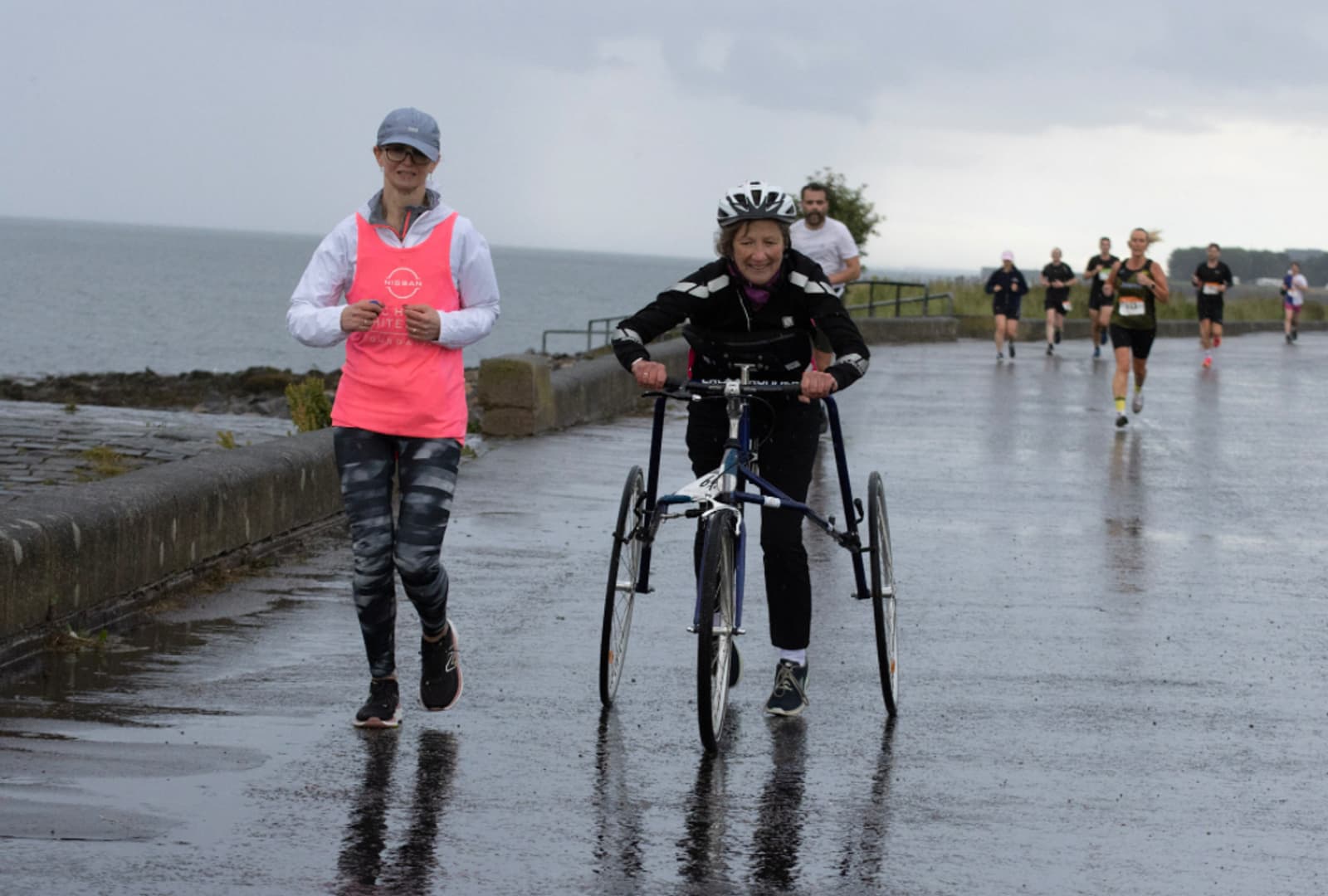 Two women participate in a race on a wet, coastal path; one walks beside a woman using a racing tricycle. Both are smiling, and other runners are visible in the background. The sky is overcast.