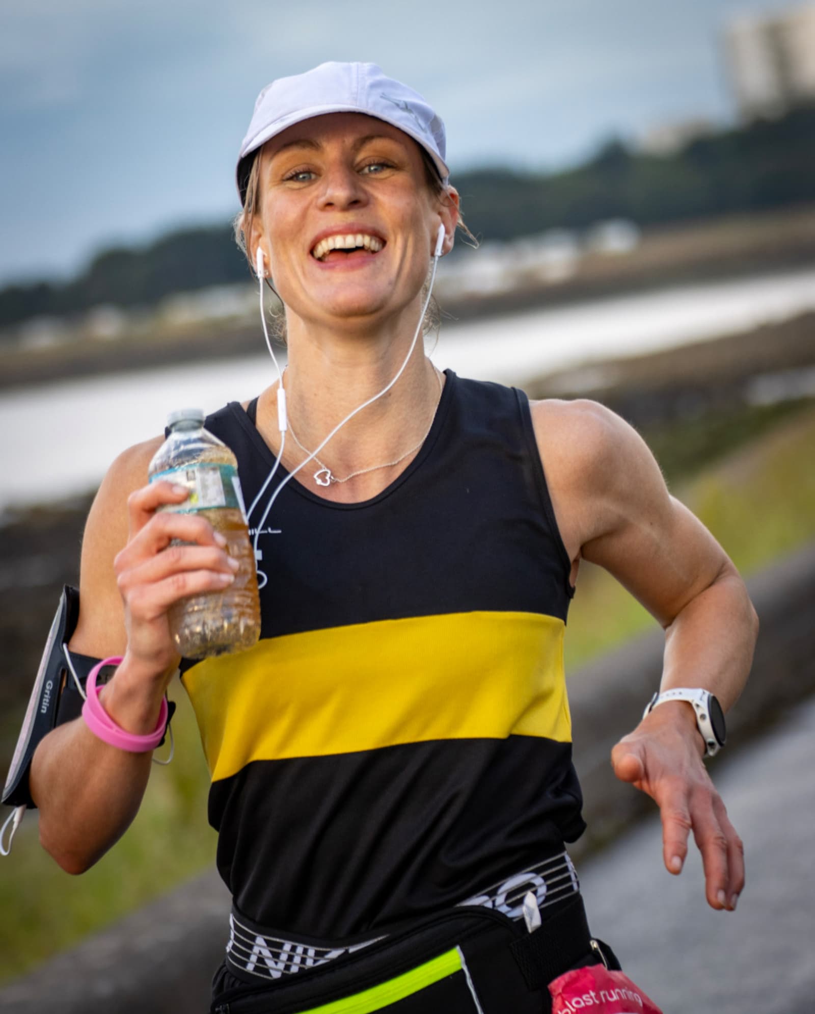 A woman in a black and yellow tank top, white cap, and earphones smiles while running outdoors, holding a bottle of water in one hand. A blurred background suggests a waterside path.