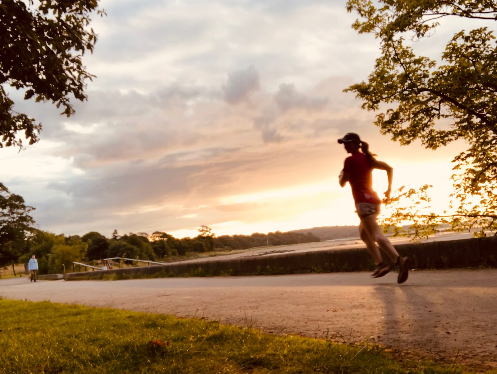 A person is jogging along a path during sunset, with the sun casting a warm glow on the surroundings. Trees frame the path on both sides, and another person walks in the distance. The sky is partly cloudy with hints of blue and orange hues.
