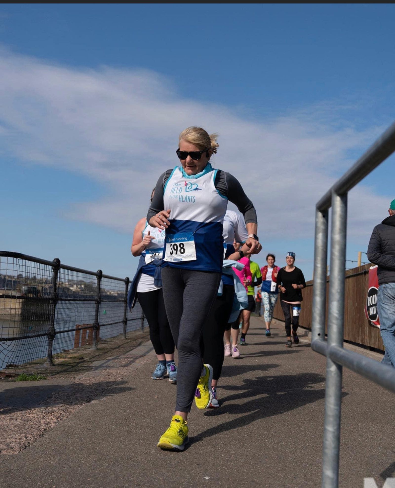 A group of runners participating in a race on a paved path next to a body of water. The lead runner, wearing a blue and white shirt, black leggings, and yellow shoes, is looking down at her watch. In the background, other runners and a clear sky are visible.