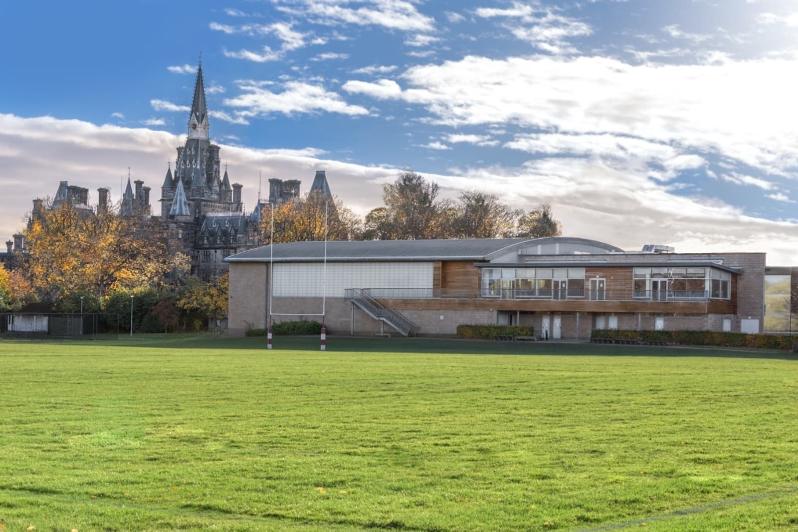 A modern building with large windows and a wooden facade is situated in front of a grassy field. Behind the contemporary structure, there's a historic, gothic-style stone building with spires, and the sky is partly cloudy with sunlight breaking through.