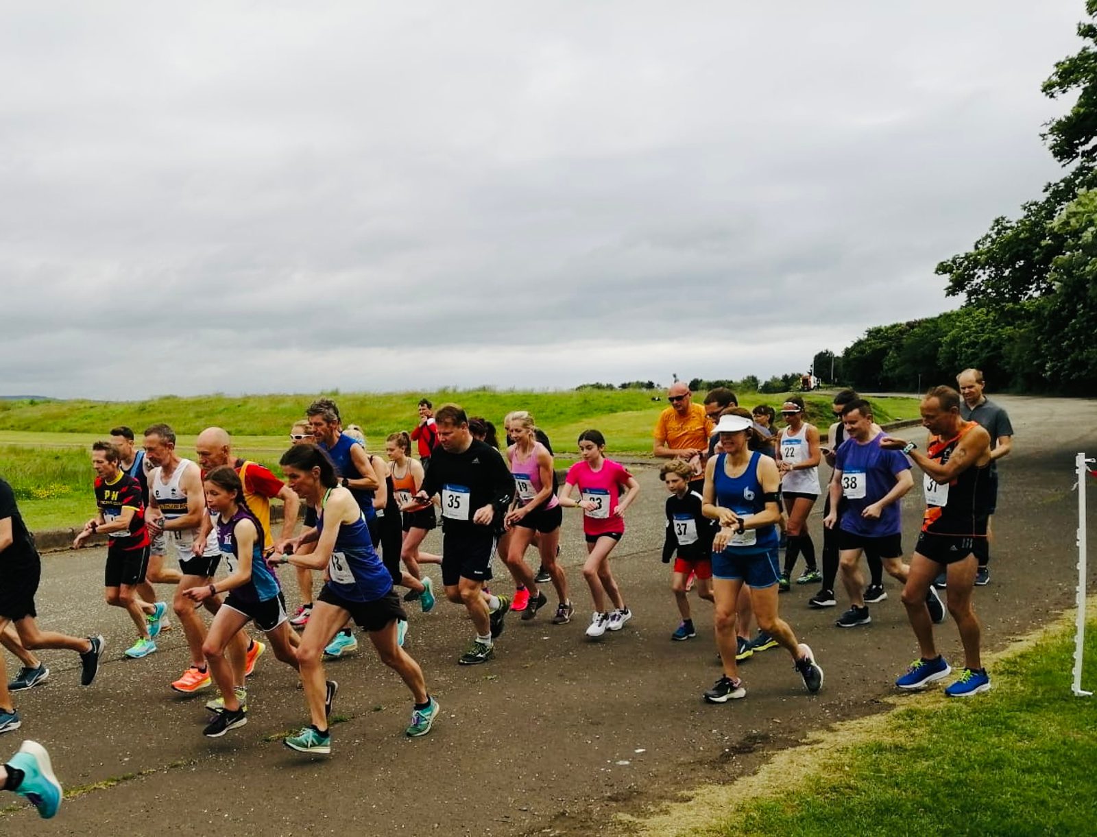 A group of runners starting a race on an outdoor path on a cloudy day. The participants, wearing various colorful athletic outfits and bib numbers, are of different ages. Green grass and trees line the pathway, and spectators are visible in the background.