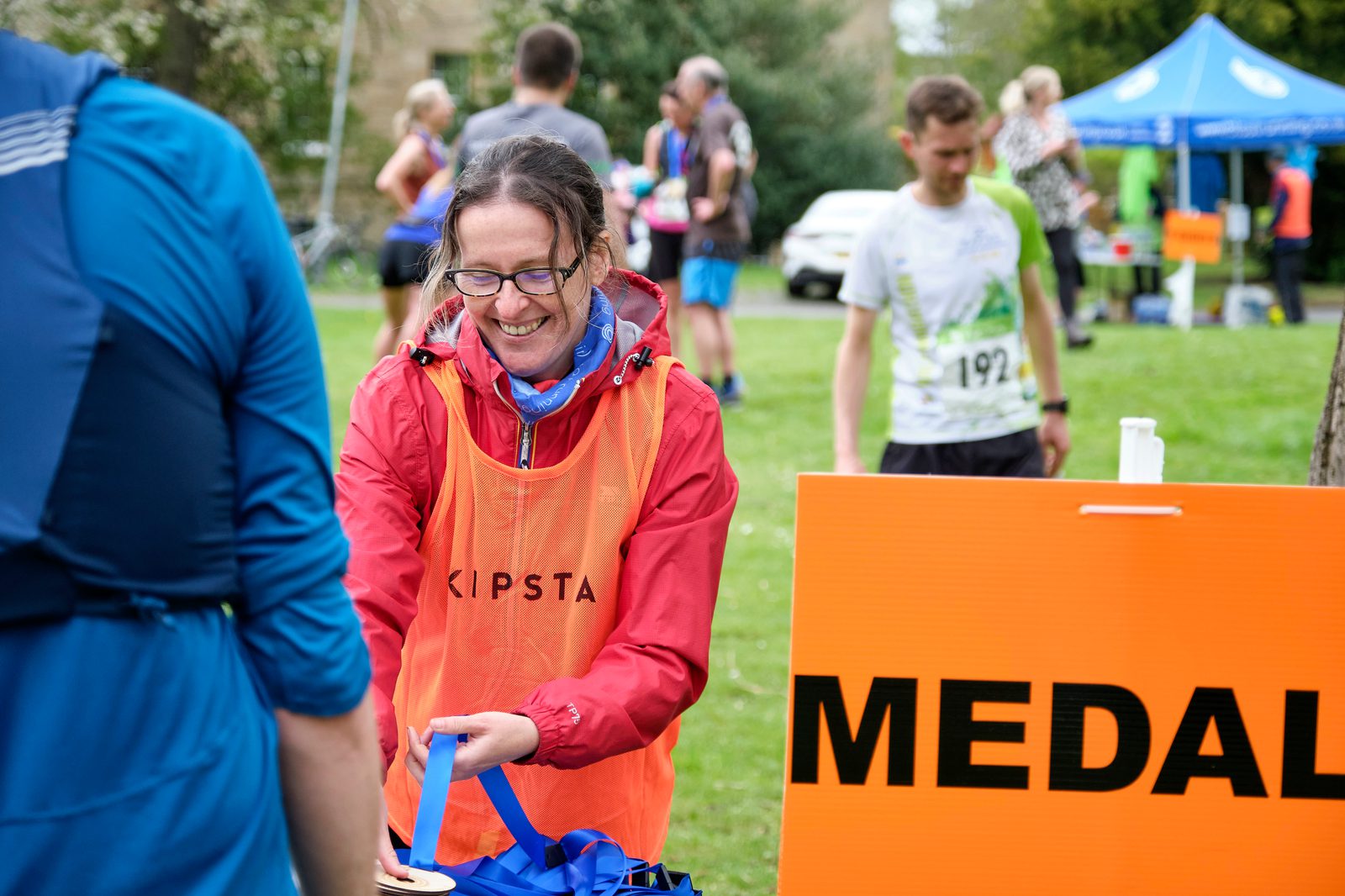 A woman wearing glasses and a red jacket with a bright orange "Kipsta" vest smiles as she handles medals at an outdoor event. A large orange sign reading "MEDALS" is visible in front of her. Other participants and a blue tent can be seen in the background.