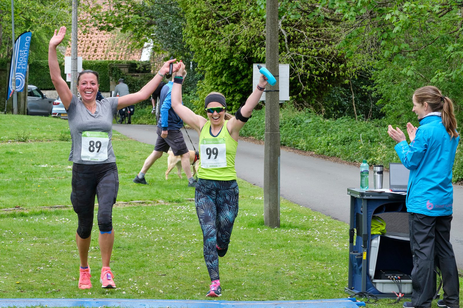 Two women cross the finish line of a race, holding hands and raising their arms in celebration. They wear race bibs numbered 89 and 99. A person in a blue jacket stands to the side, clapping. The background features greenery and a few other participants.
