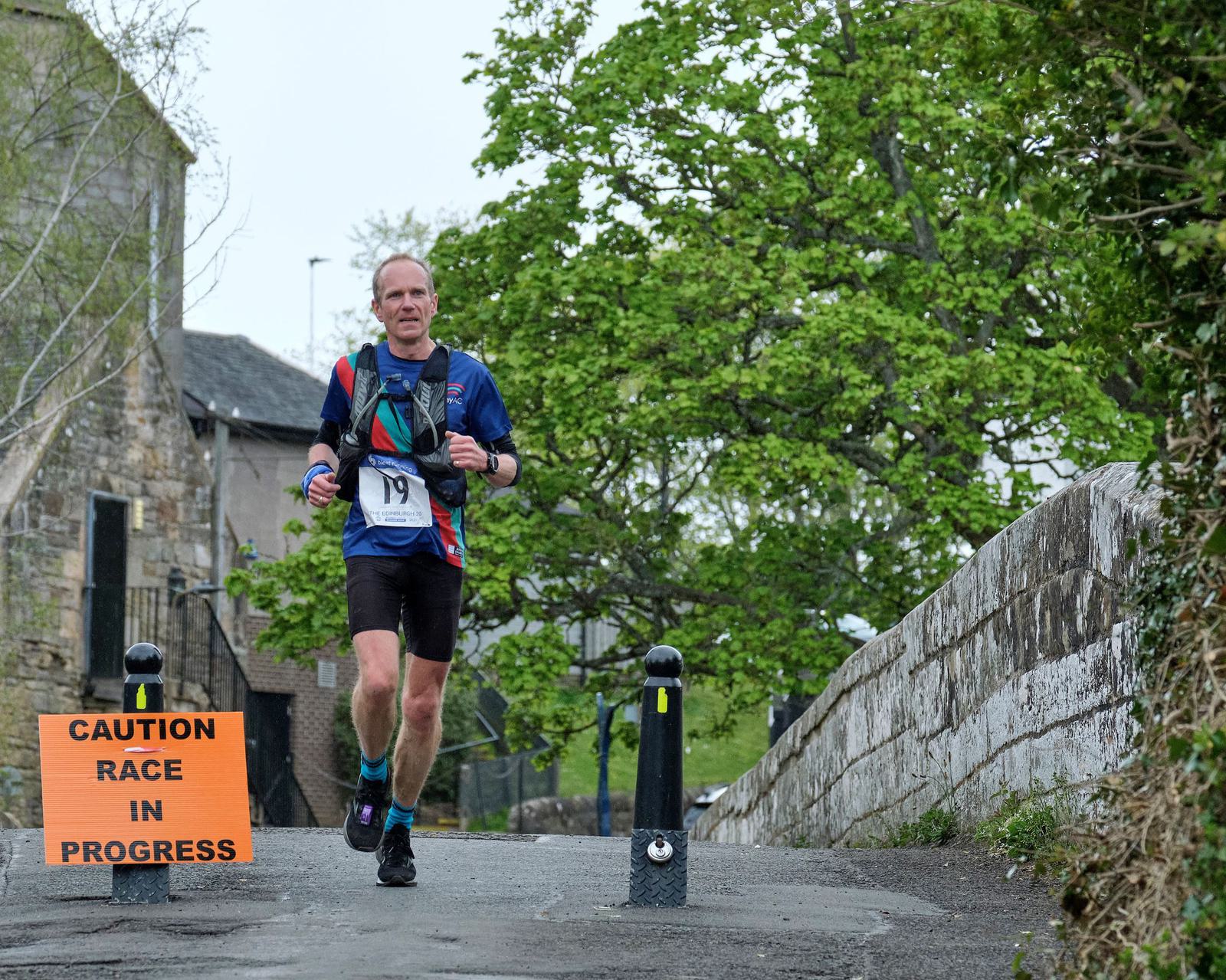 A runner in a blue and red shirt, bib number 19, is running on a paved path next to a brick wall. A bright yellow sign on the left reads, "CAUTION RACE IN PROGRESS." Trees and buildings are visible in the background.