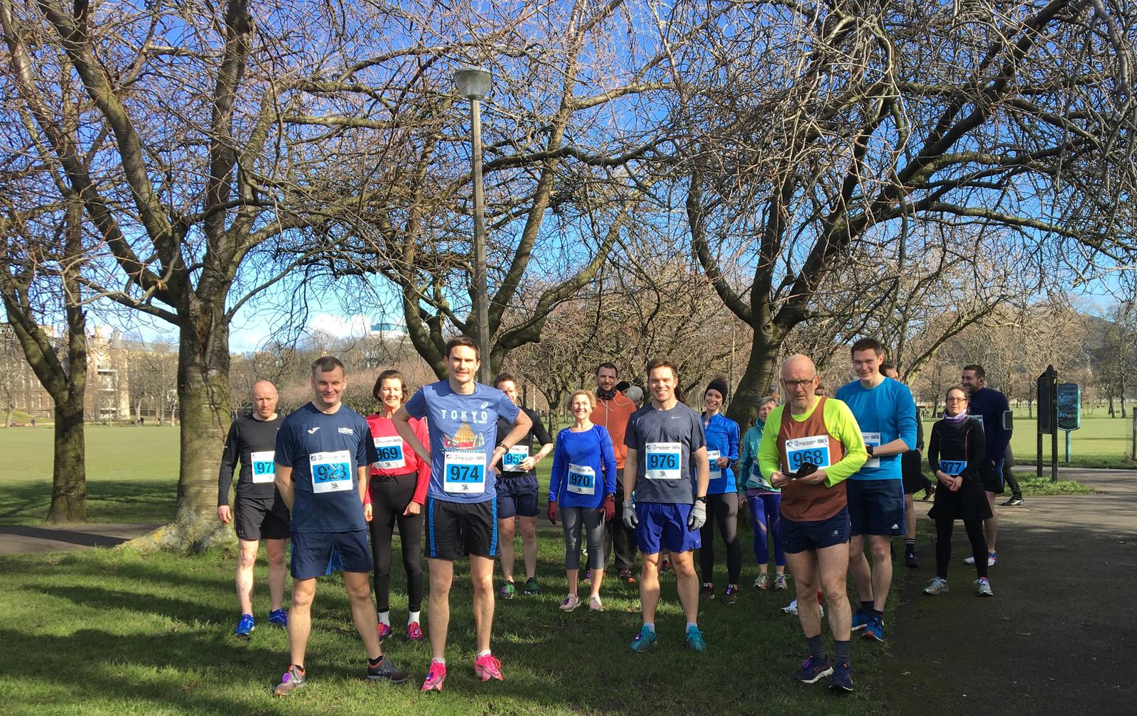 A group of runners wearing numbered bibs are gathered in a park on a clear day. The runners are dressed in athletic gear, and the trees around them are bare, indicating it might be early spring or late winter. Some are smiling and appear ready for a race or group run.