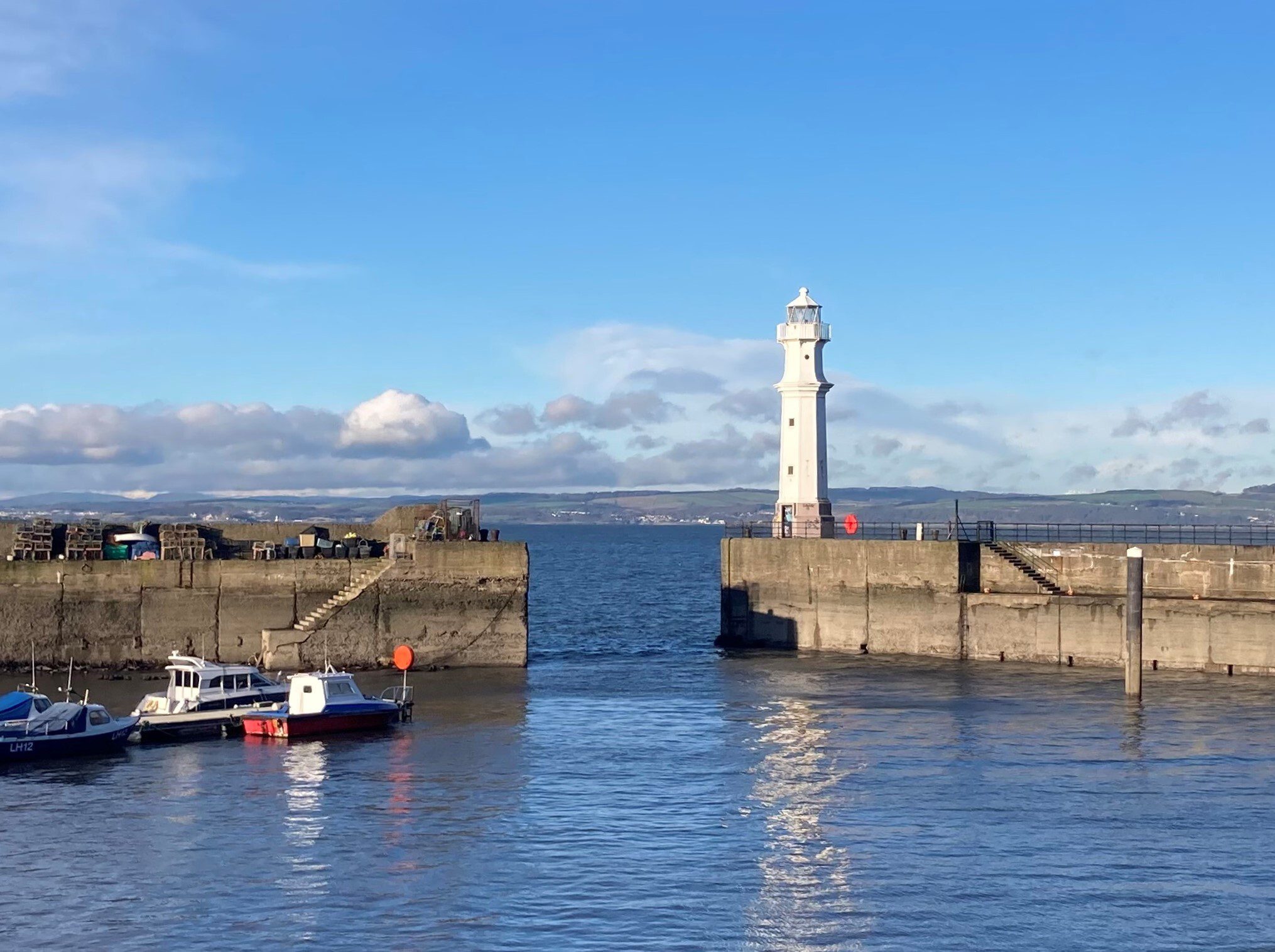 A serene waterfront scene featuring a white lighthouse standing at the end of a concrete jetty. Two small boats are moored in the calm blue water nearby. The sky is clear with scattered clouds, and distant land is visible on the horizon.
