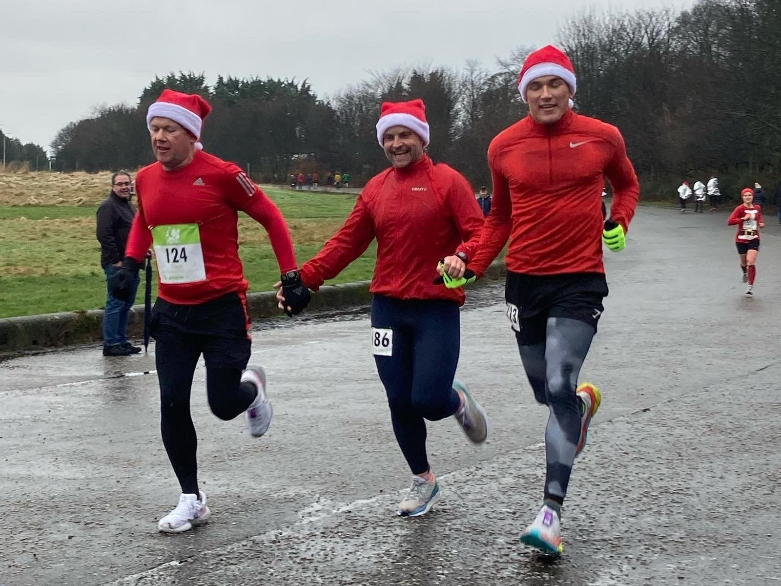 Three runners, dressed in red shirts and Santa hats, run hand in hand during a race. The runner in the middle has bib number 86, while the runner on the left has bib number 124. The background shows a park with sporadic onlookers and more runners in the distance.