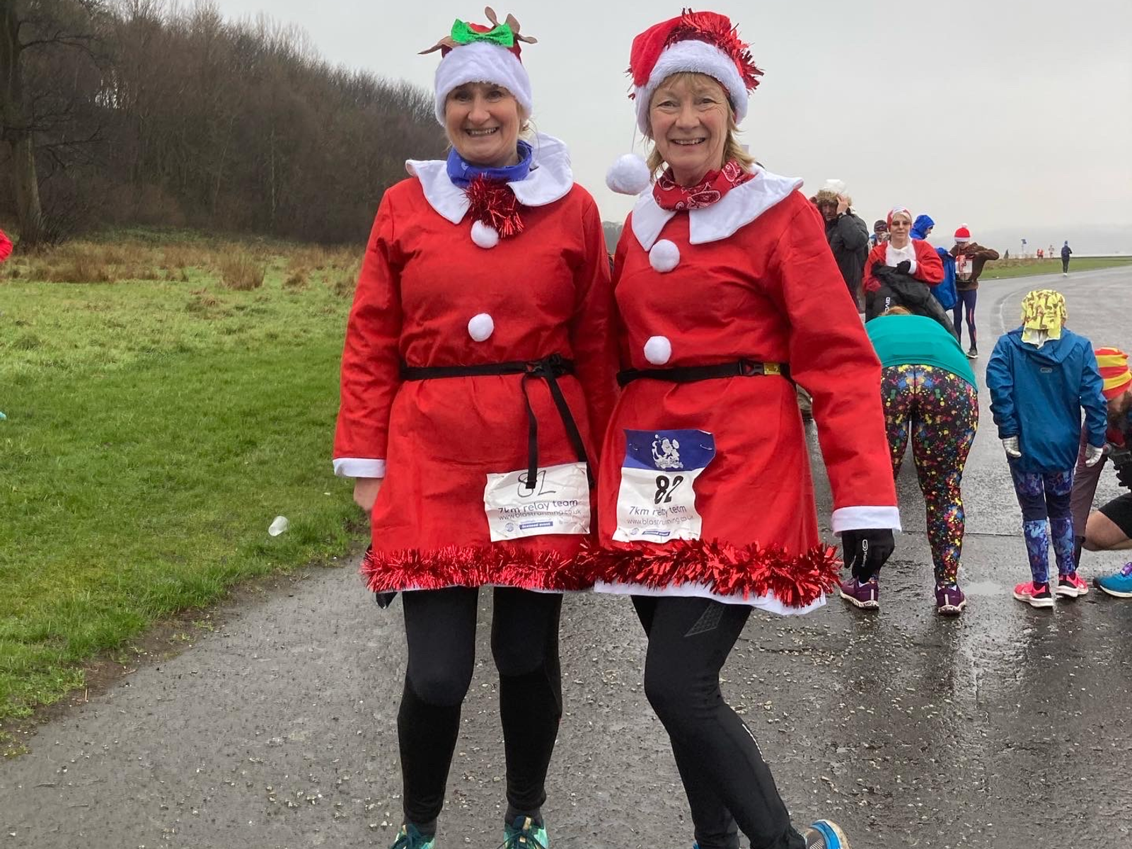 Two women dressed in festive Santa outfits stand on a paved path with a grassy area in the background. Both are smiling and wearing Christmas hats, one with a Santa hat and the other with reindeer antlers. Other people in colorful attire are gathered in the background.