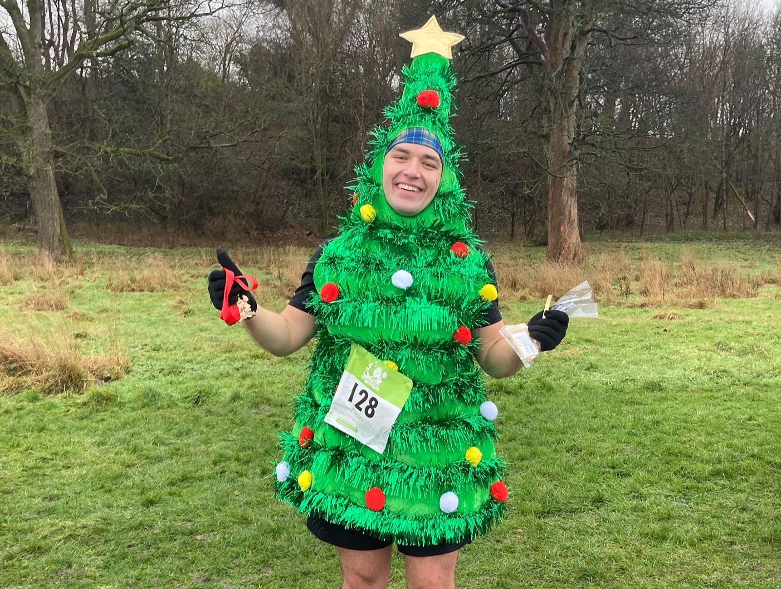 A person stands outdoors on a grassy area surrounded by trees, dressed in a Christmas tree costume adorned with tinsel, ornaments, and a star on top. They are smiling, holding a medal in one hand and a small bag in the other. They wear a race number, 128, on their chest.