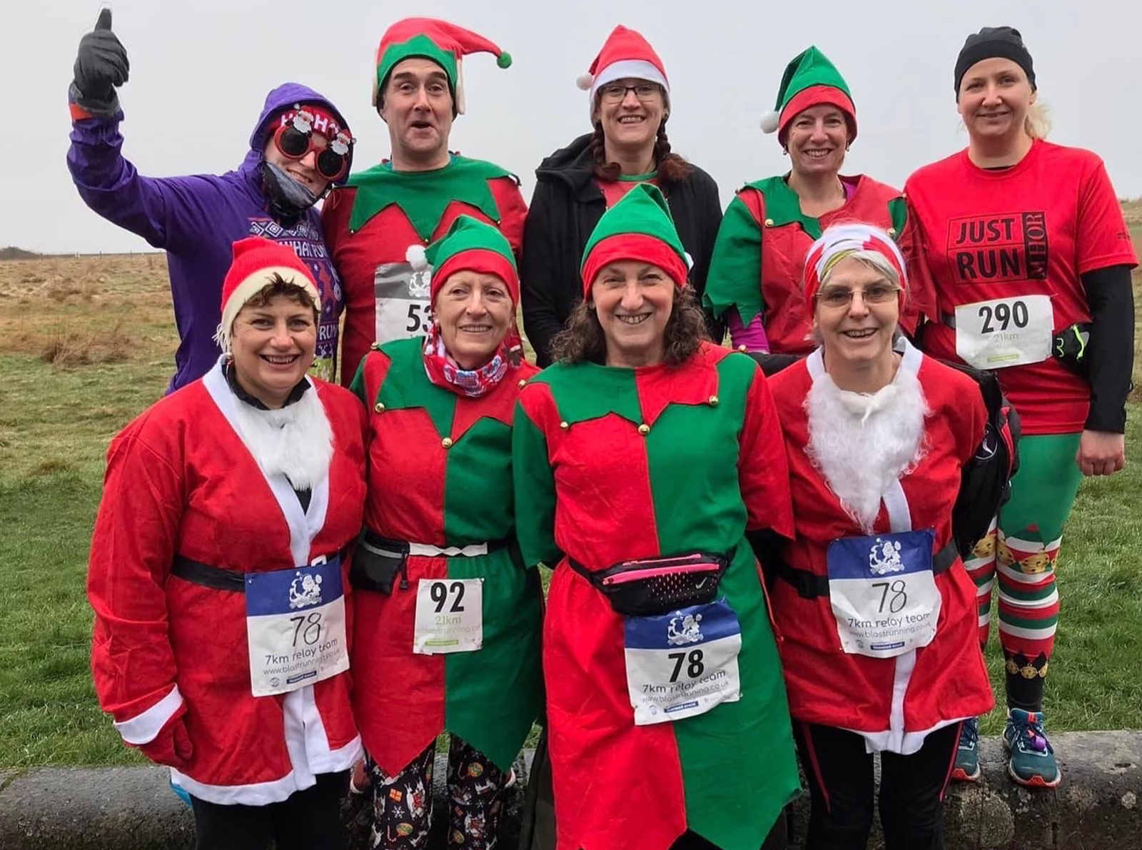 A group of nine runners dressed in festive holiday costumes, including Santa and elf outfits, smile for a group photo. They are outdoors on a grassy area with race bibs pinned to their clothes, indicating participation in a themed race or fun run.
