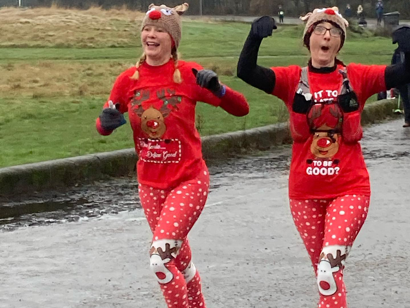 Two people are running on a gray path, wearing festive red outfits with reindeer designs and reindeer antler hats. They appear cheerful and are smiling. One is wearing glasses. The background includes a grassy area.