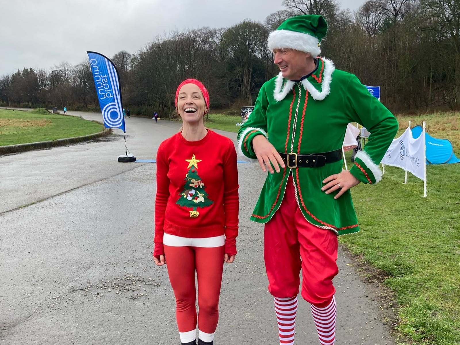 A smiling person dressed in a red Christmas tree outfit stands next to a tall person dressed as a green elf with red shorts and a Santa hat. They are outdoors on a rainy day with parkland and event flags in the background.