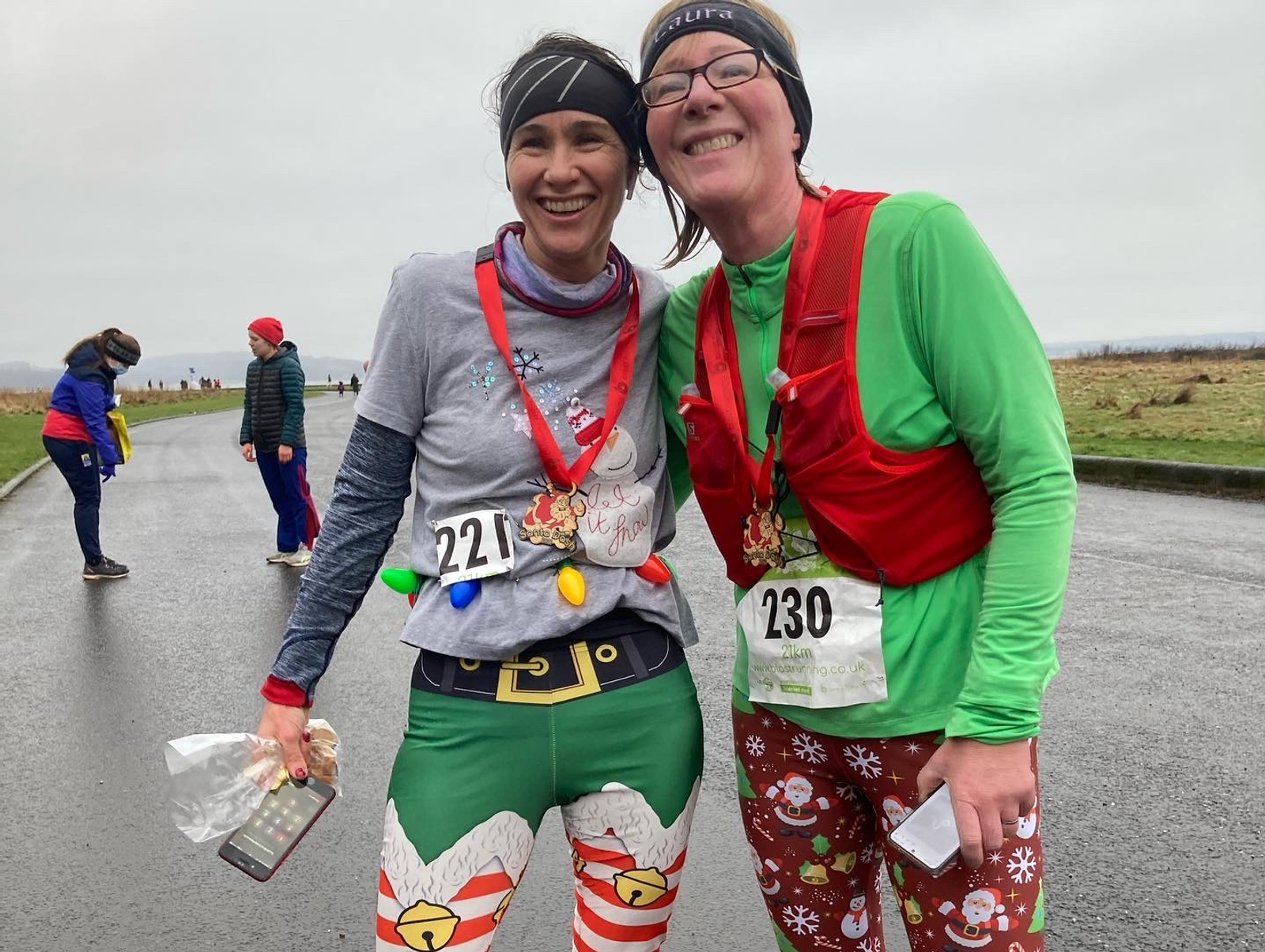 Two runners in festive outfits smile for the camera after a race. The runner on the left wears a grey shirt with a Santa design, elf pants, and a race number 221. The runner on the right wears a red vest, green shirt, festive pants, and a race number 230.