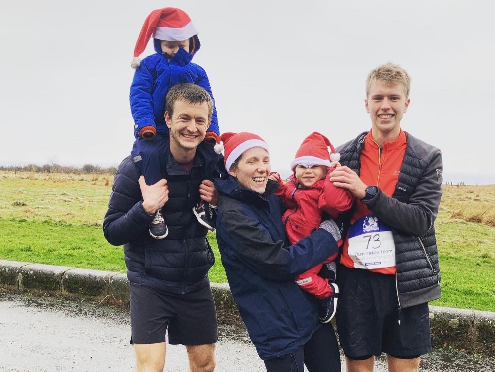 A group of four people smiling outdoors with two children. One adult is carrying a child on their shoulders while another adult is holding a child dressed in festive red clothing. Both children and one adult wear Santa hats. The number "73" is visible on a race bib.