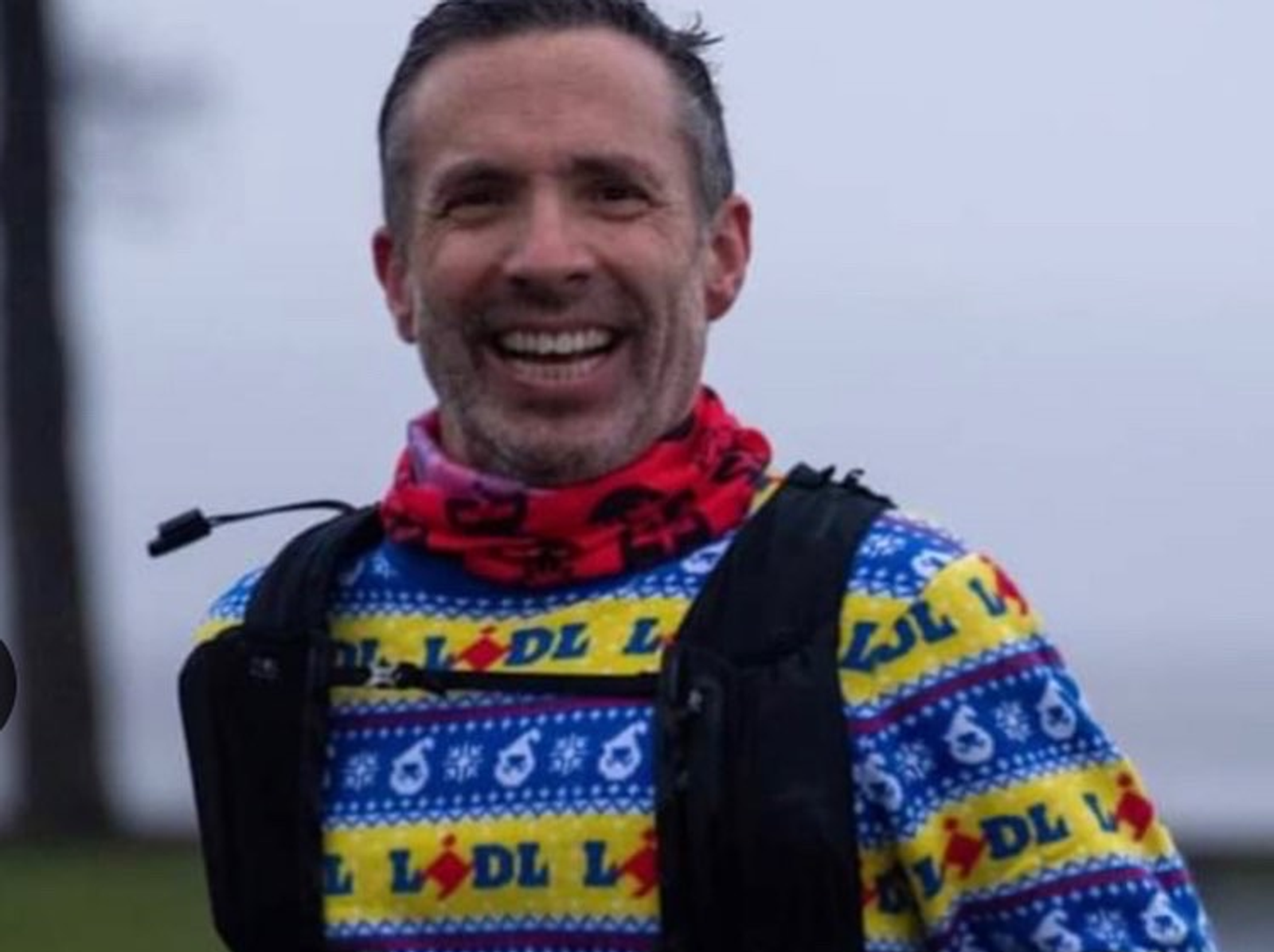 A man smiles broadly while wearing a colorful sweater with the word "Lidl" and snowflake patterns on it. He has a red neck gaiter and a black running vest. The background is slightly blurred, hinting at an outdoor setting.