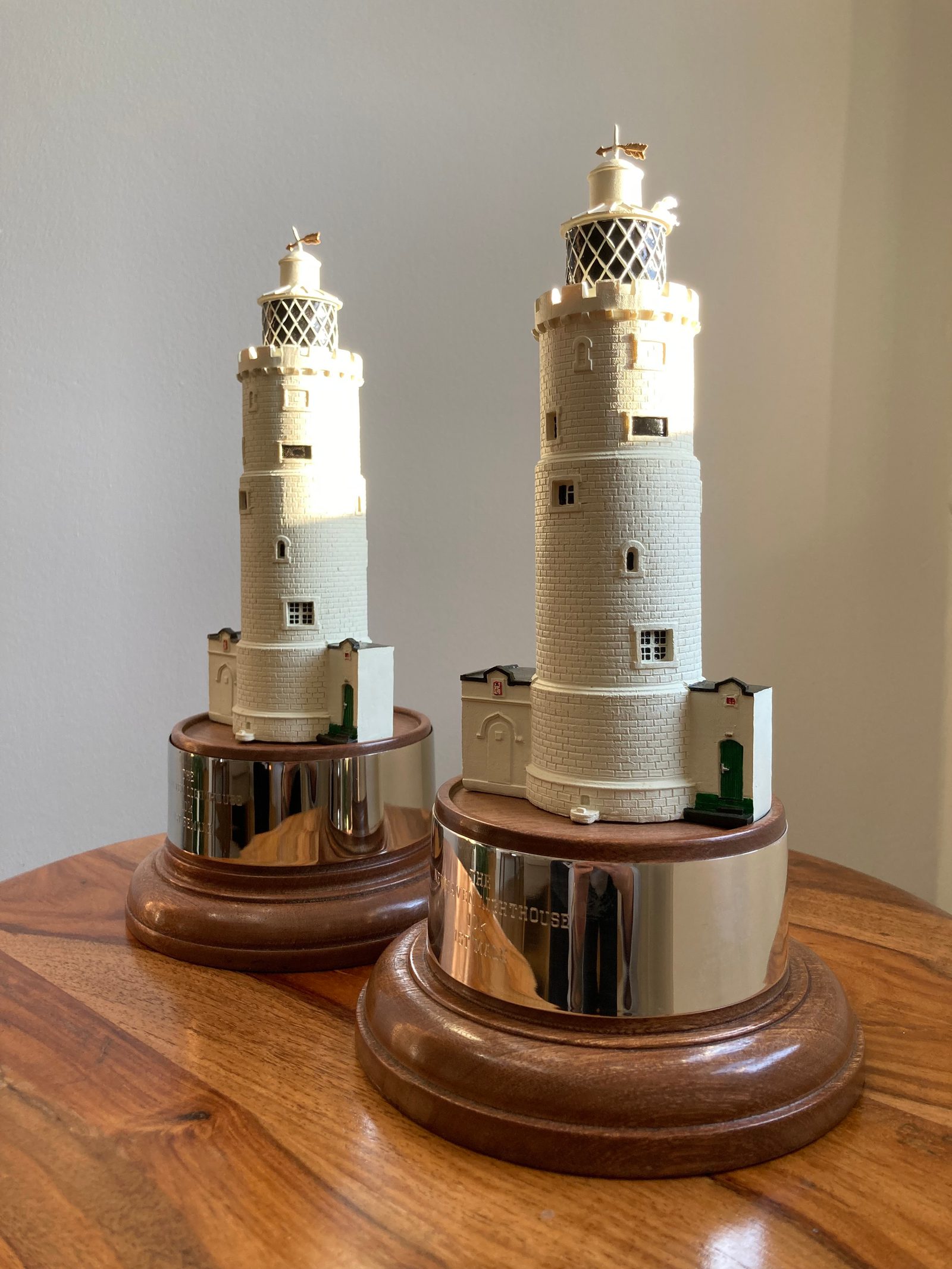 Two detailed lighthouse models sit on a wooden table. The lighthouses are mounted on round wooden bases with metal plaques. Each model features a white cylindrical tower with small windows and a balcony near the top. Sunlight highlights the models.