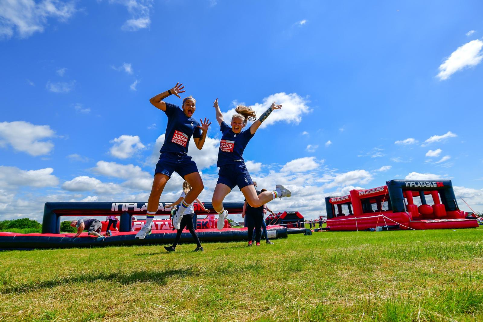 Two people wearing race bibs jump joyfully in a grassy field with inflatable obstacle course structures in the background, under a blue sky with scattered clouds.