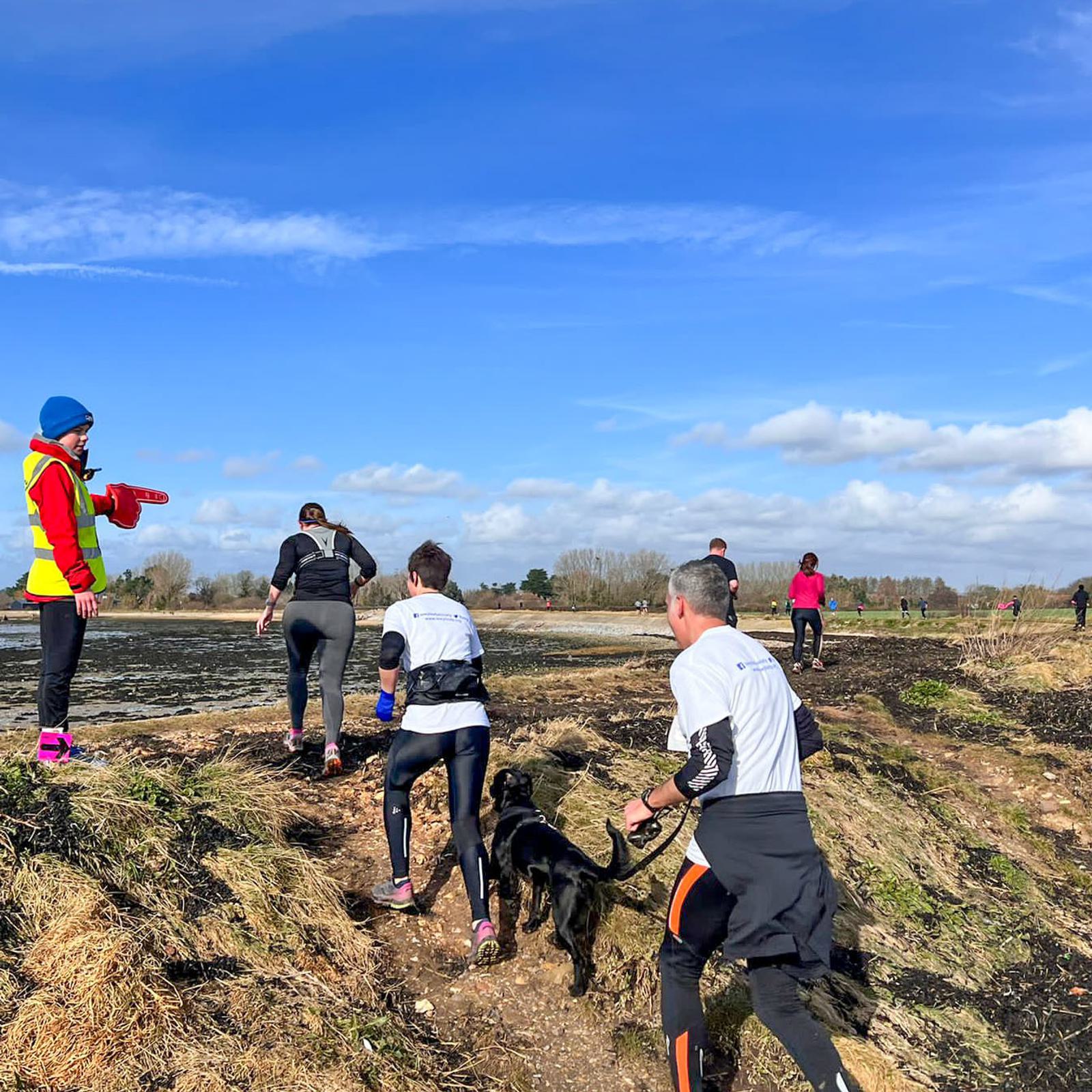 People jogging along a coastal path under a clear blue sky. A person in a yellow vest gestures with a megaphone. A man holding a dog's leash is in the foreground. Other runners are ahead, with some vegetation and sparse clouds in the background.