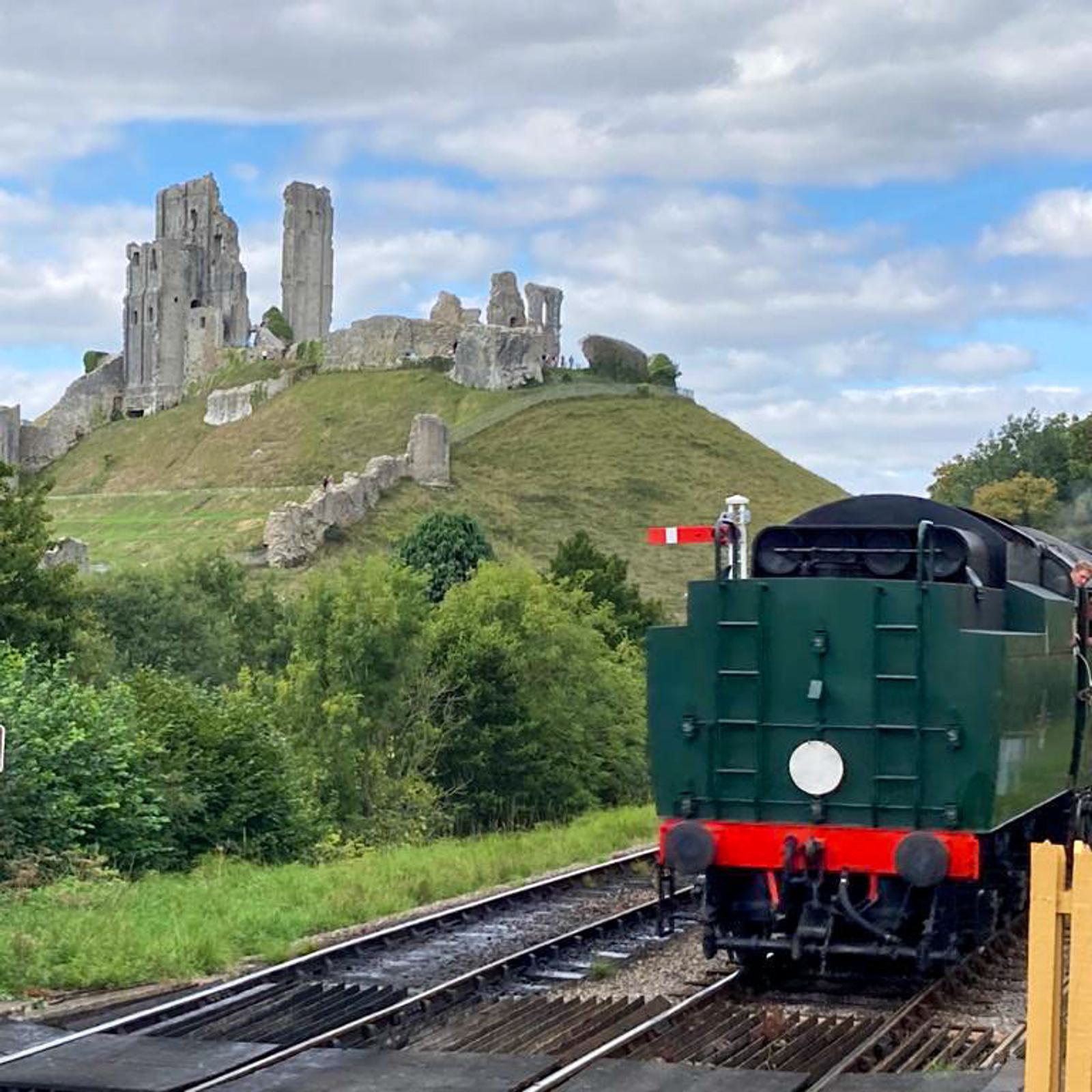 A green steam train travels on tracks with the ruins of a castle on a hill in the background. The sky is partly cloudy, and lush greenery surrounds the scene.