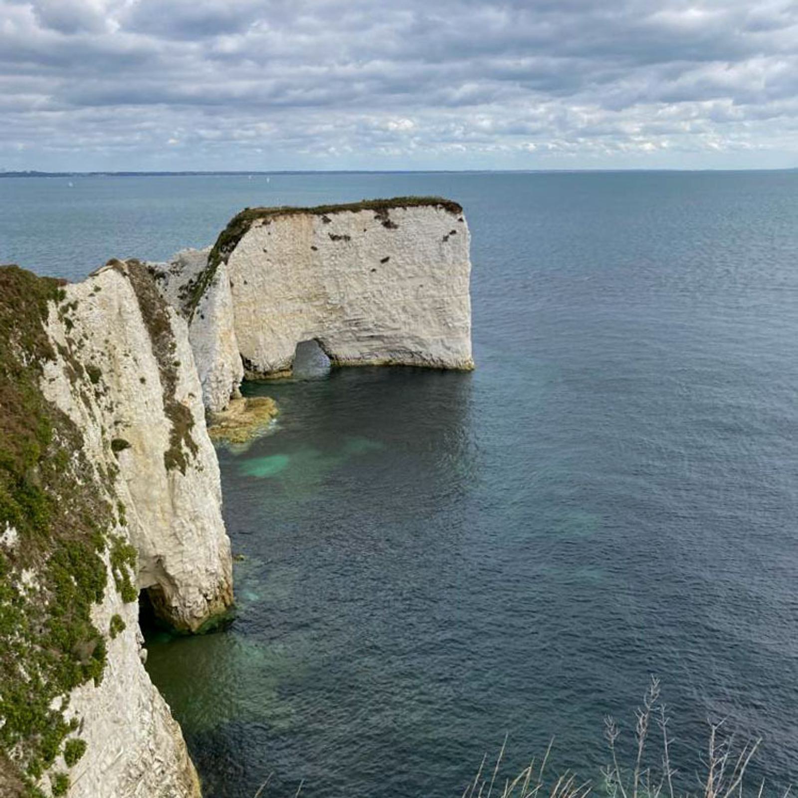Cliffs along the coastline with a prominent rock formation jutting into the sea under a cloudy sky. The ocean is calm with spots of deeper blue near the rocks. Sparse vegetation covers parts of the cliffs.