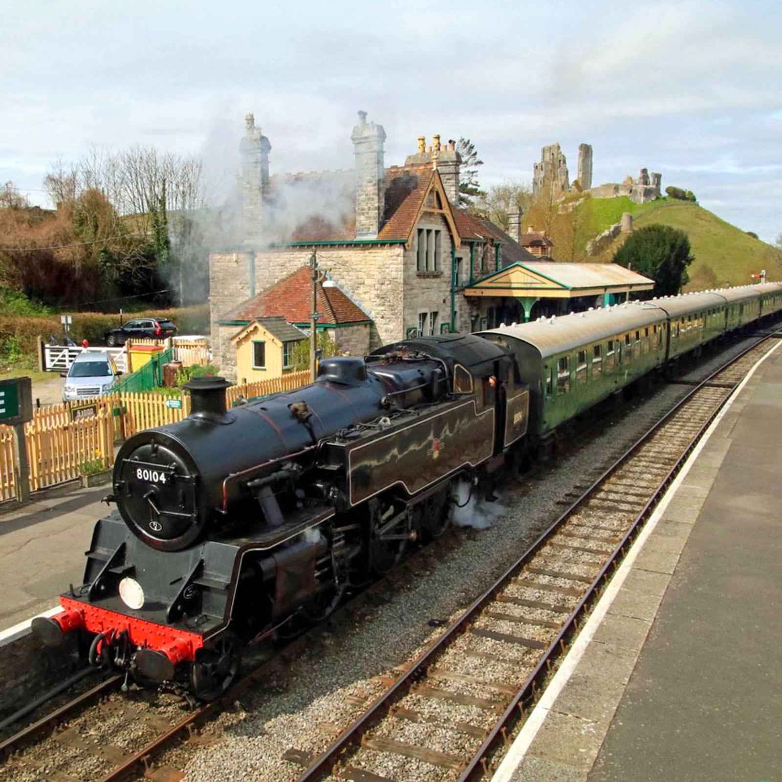 A vintage black steam train with a green passenger carriage passes through a quaint rural station. Stone buildings and a castle on a hill are in the background. The train emits white steam under a partly cloudy sky.