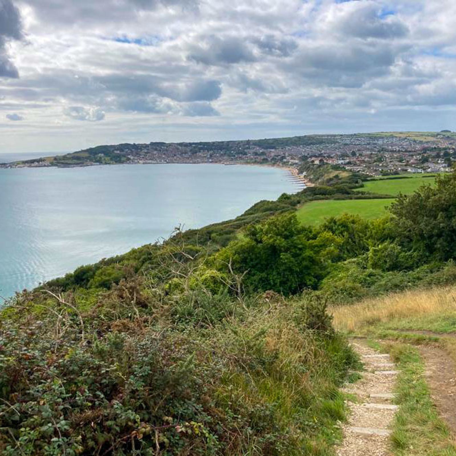A coastal landscape with a narrow dirt path leading down a hillside, surrounded by greenery. The view overlooks a curved bay with calm water and a town in the distance under a cloudy sky.