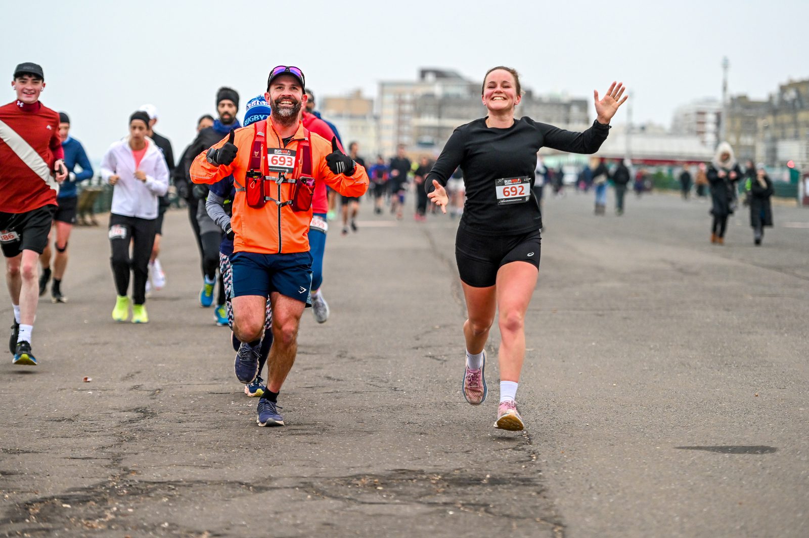 Two smiling runners, a man in an orange jacket and a woman in black, participate in a race on a city street, with other runners and buildings visible in the background.