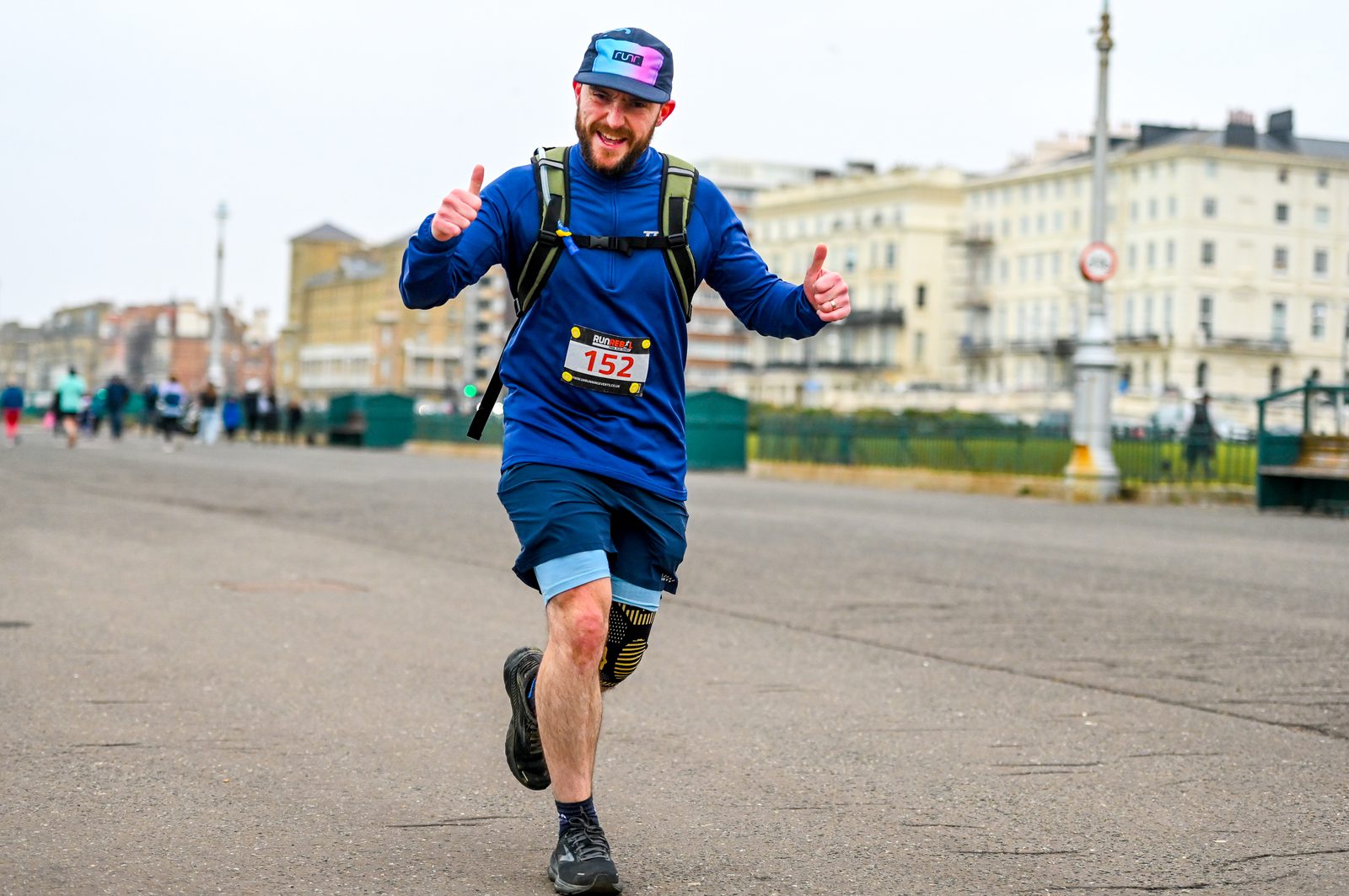 A smiling runner in a blue outfit and cap, wearing a race bib numbered 152, gives two thumbs up while running on a street lined with buildings during a race event.