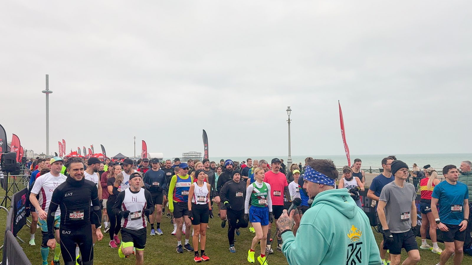 A large group of runners gathers at the start line on a grassy area near the ocean, some wearing race bibs and athletic gear, while a man in a mint hoodie addresses them under a cloudy sky.