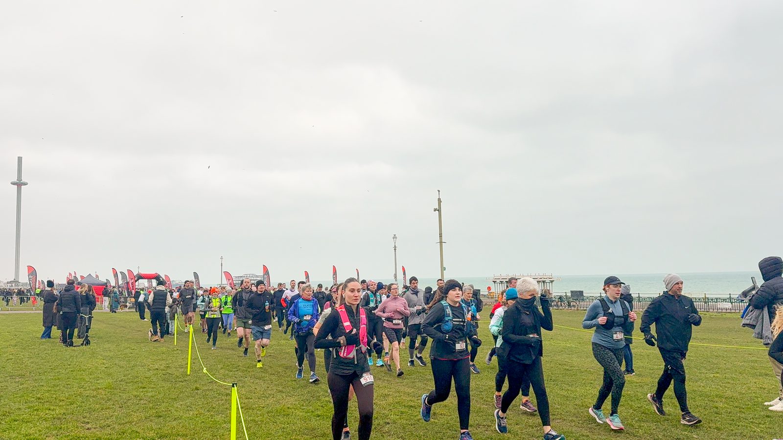 A group of people in athletic clothing are running on grass near the sea, under a cloudy sky, with red flags and event markers in the background.