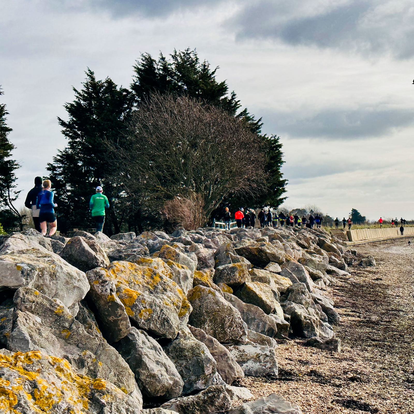 A group of people jogs along a path beside a rocky shoreline. Leafless trees are visible in the background under a cloudy sky. The scene conveys a sense of outdoor activity.