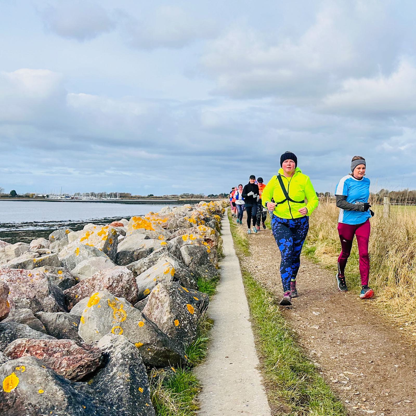 A group of people jogging on a narrow path by the sea. Large rocks are on the left and grassy areas on the right. The sky is cloudy. The lead runner is wearing a bright yellow jacket, and others follow behind.