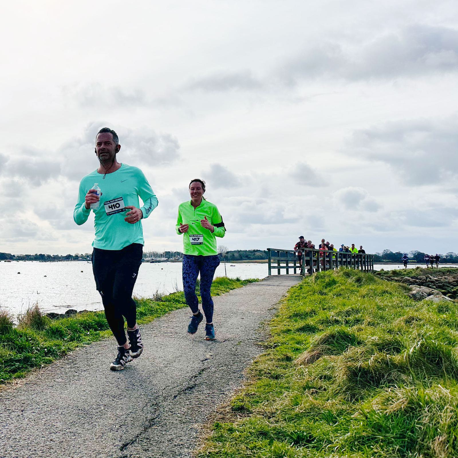 Two runners in bright shirts jog along a paved path by a body of water. They are part of an organized event, indicated by bibs on their shirts. The sky is cloudy, and there are more participants in the distance near a bridge.