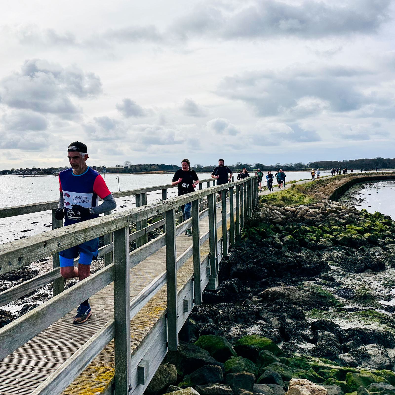 Runners participating in a race cross a wooden bridge over a rocky shoreline. The cloudy sky looms overhead as participants, wearing numbered race bibs, proceed along the path. The scene conveys a sense of determination and outdoor activity.