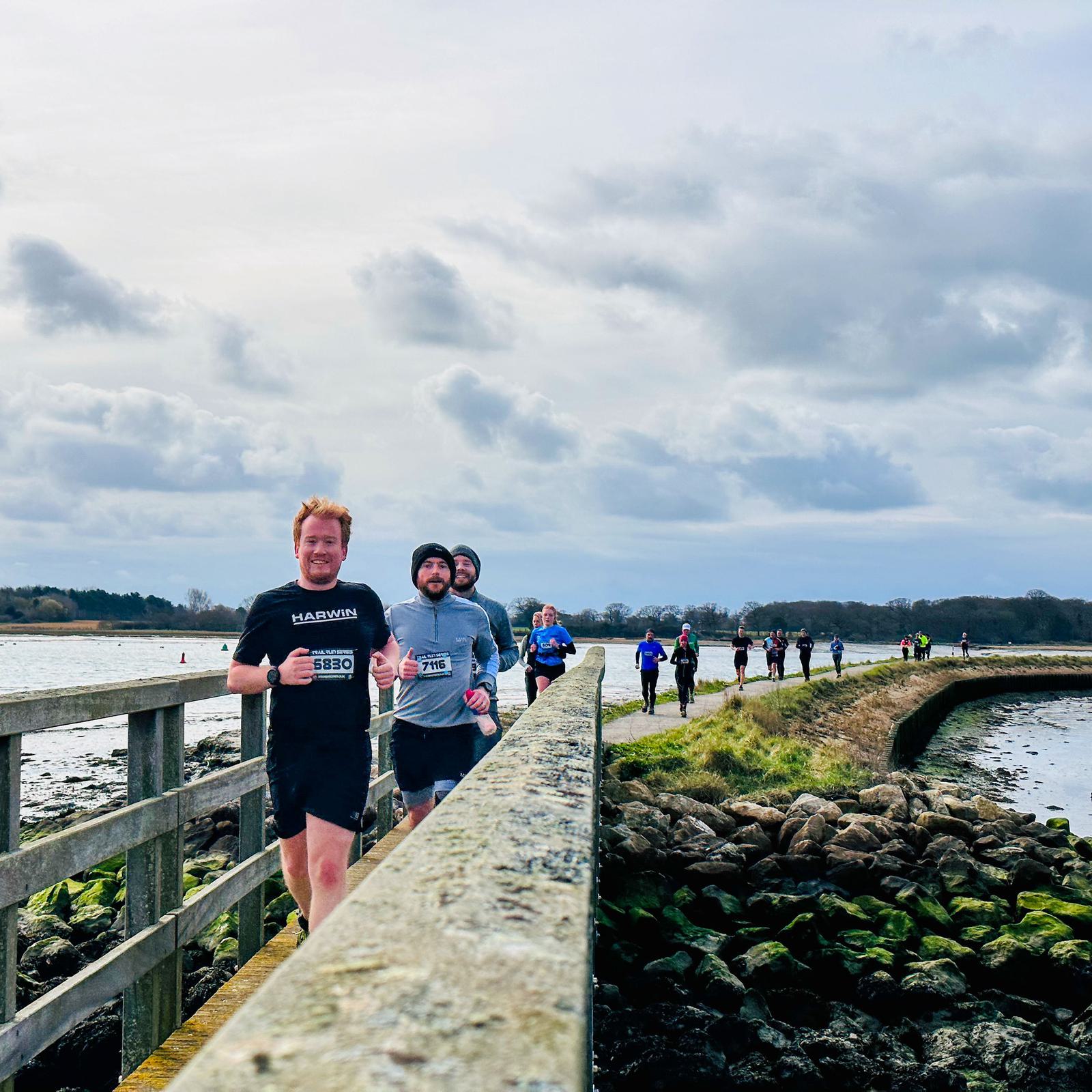 A group of runners participating in a race along a coastal path. The path is bordered by water on one side and rocky terrain on the other. The sky is cloudy, and the runners wear various athletic outfits.